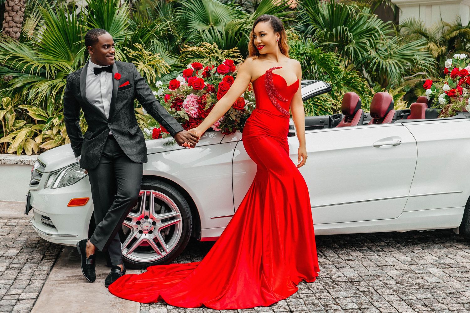 Glamorous couple poses by white convertible car, featuring a flowing red mermaid gown.