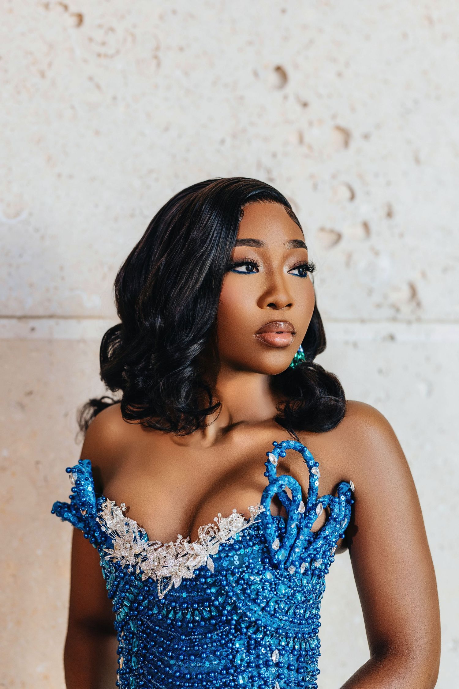 Glamorous woman in sparkling blue evening gown with sequins and dramatic curled hairstyle against textured wall.