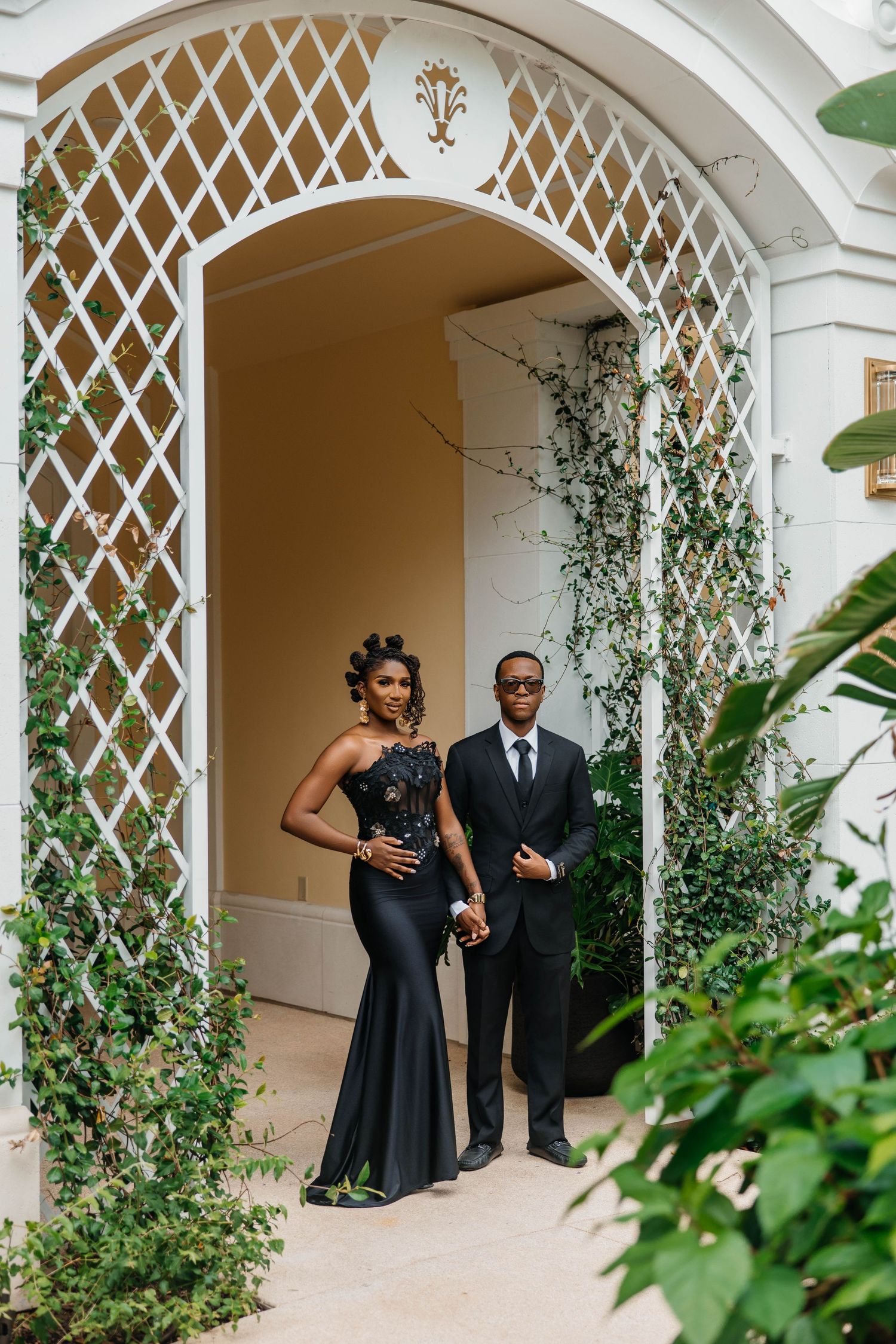 Elegant couple in black formal attire posing under a white lattice arch surrounded by lush green foliage.