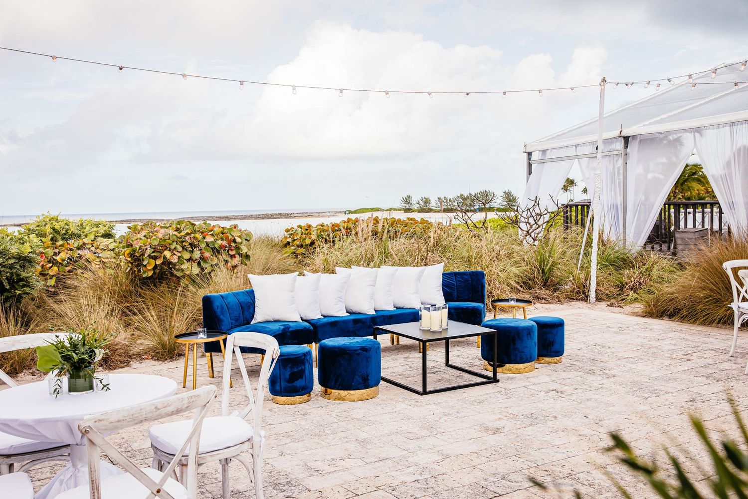 Elegant outdoor lounge with royal blue velvet sofas and gold ottomans on a sandy patio overlooking a coastal waterfront.