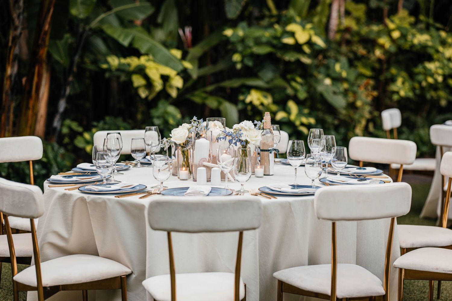 Elegant outdoor wedding reception table with white linens, gold chairs, floral centerpieces, and glassware set in a lush garden.