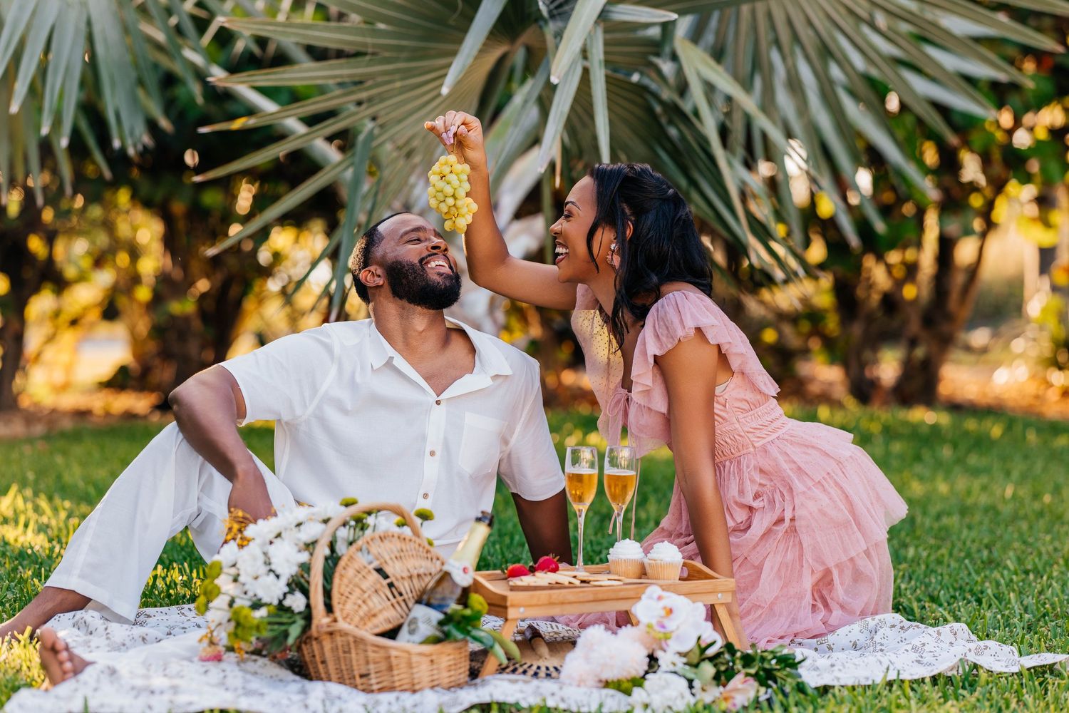 A romantic couple enjoys a sunset picnic on a blanket with cocktails and snacks under palm trees.