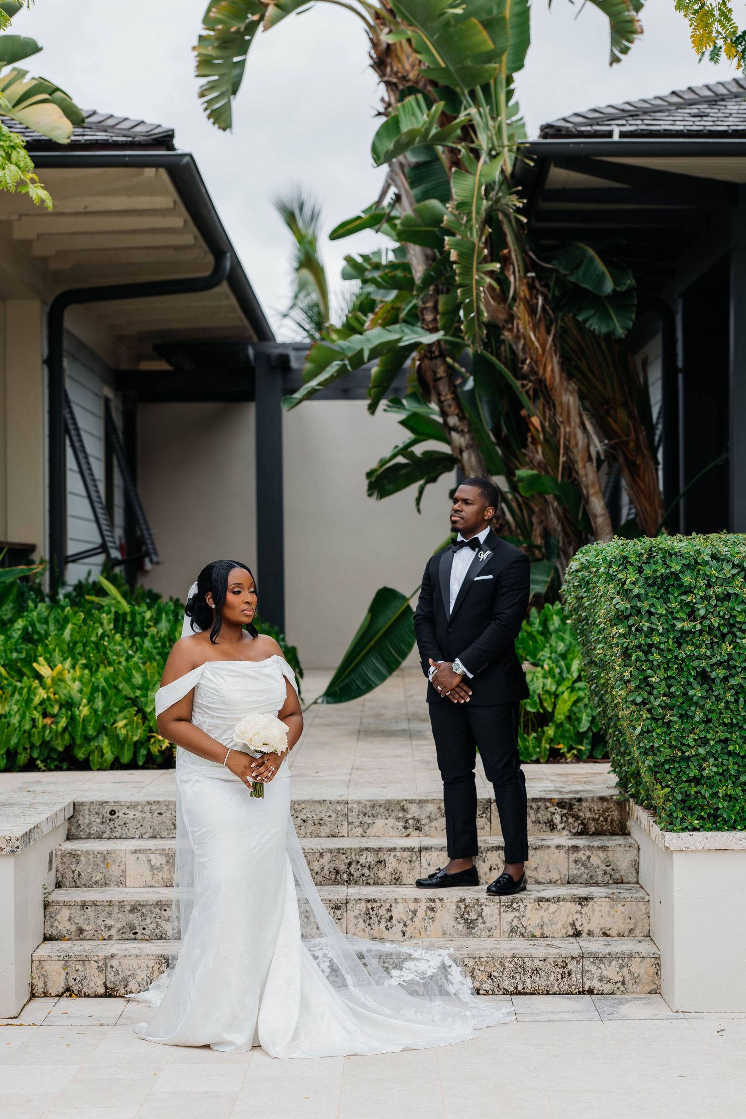Bride in off-shoulder white gown and groom in black tuxedo pose on stone steps surrounded by tropical greenery.