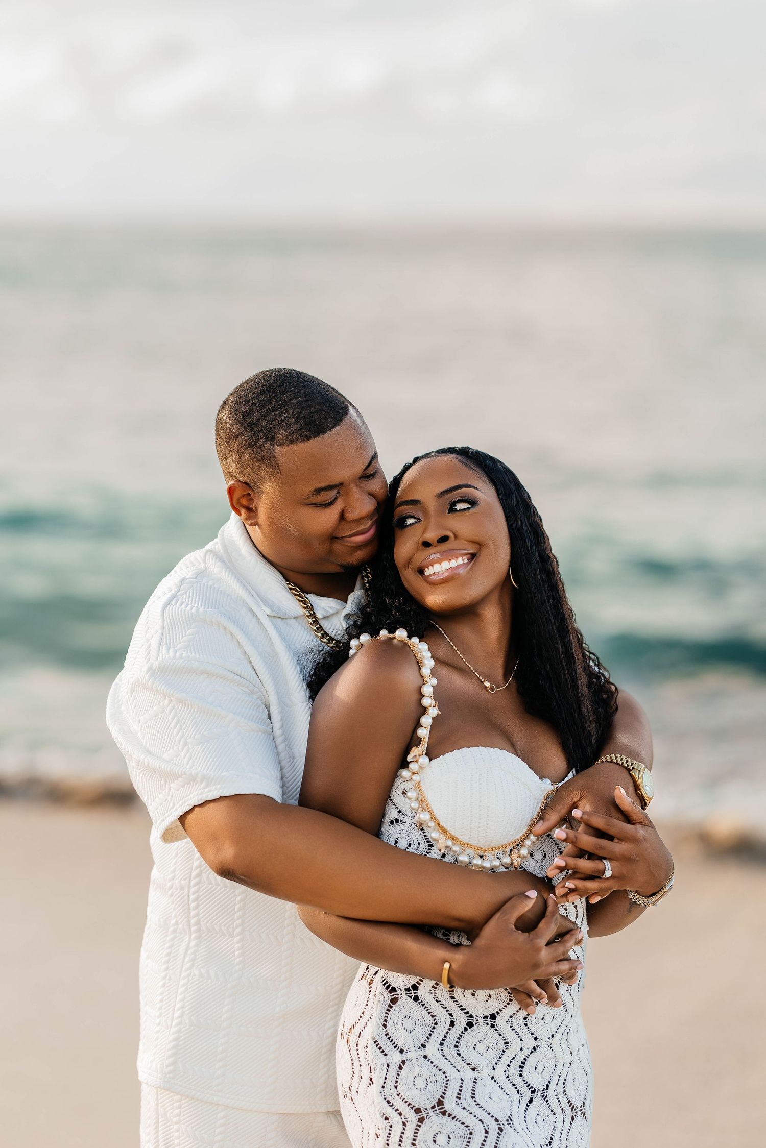 A romantic couple shares intimate moments together on the beach wearing white outfits at sunset.