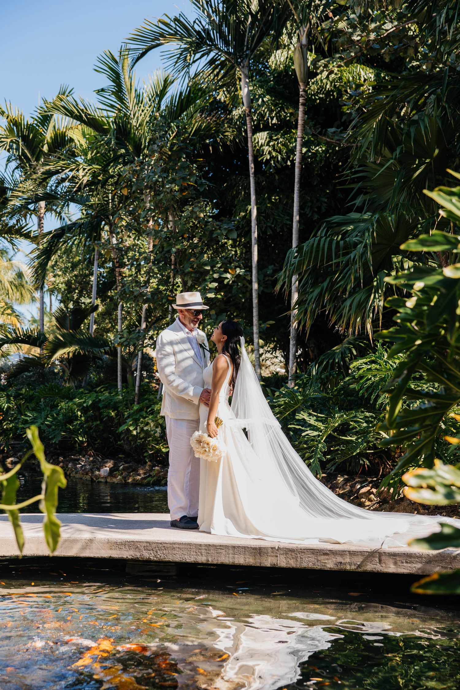 A couple in wedding attire stands together in a lush tropical garden setting with palm trees and greenery.