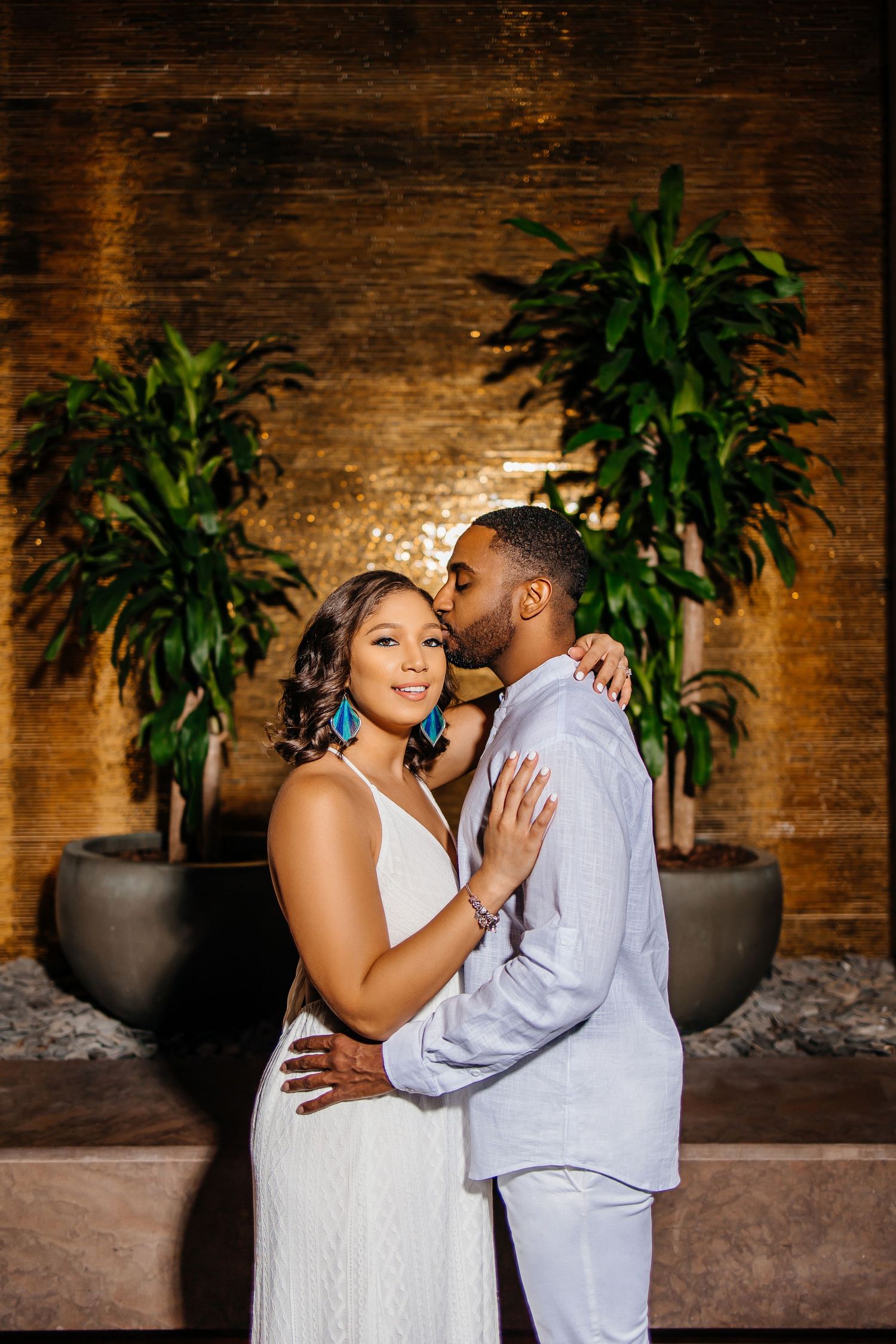 Indoor portrait against warm brick wall with decorative potted plants.