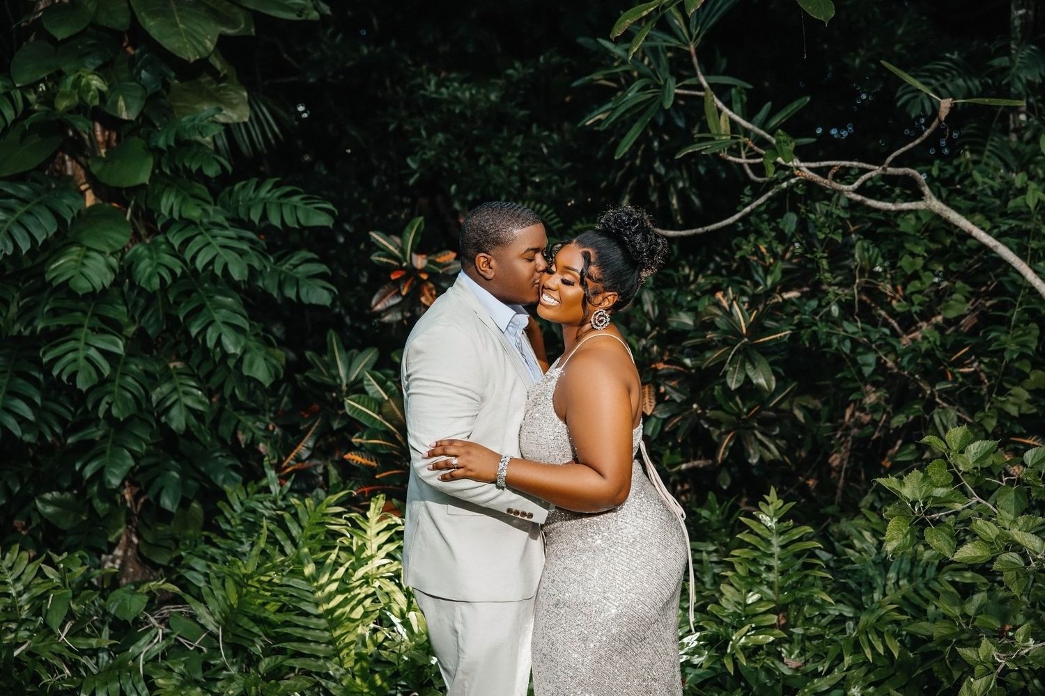 Couple embraces in tropical garden wearing white wedding attire.