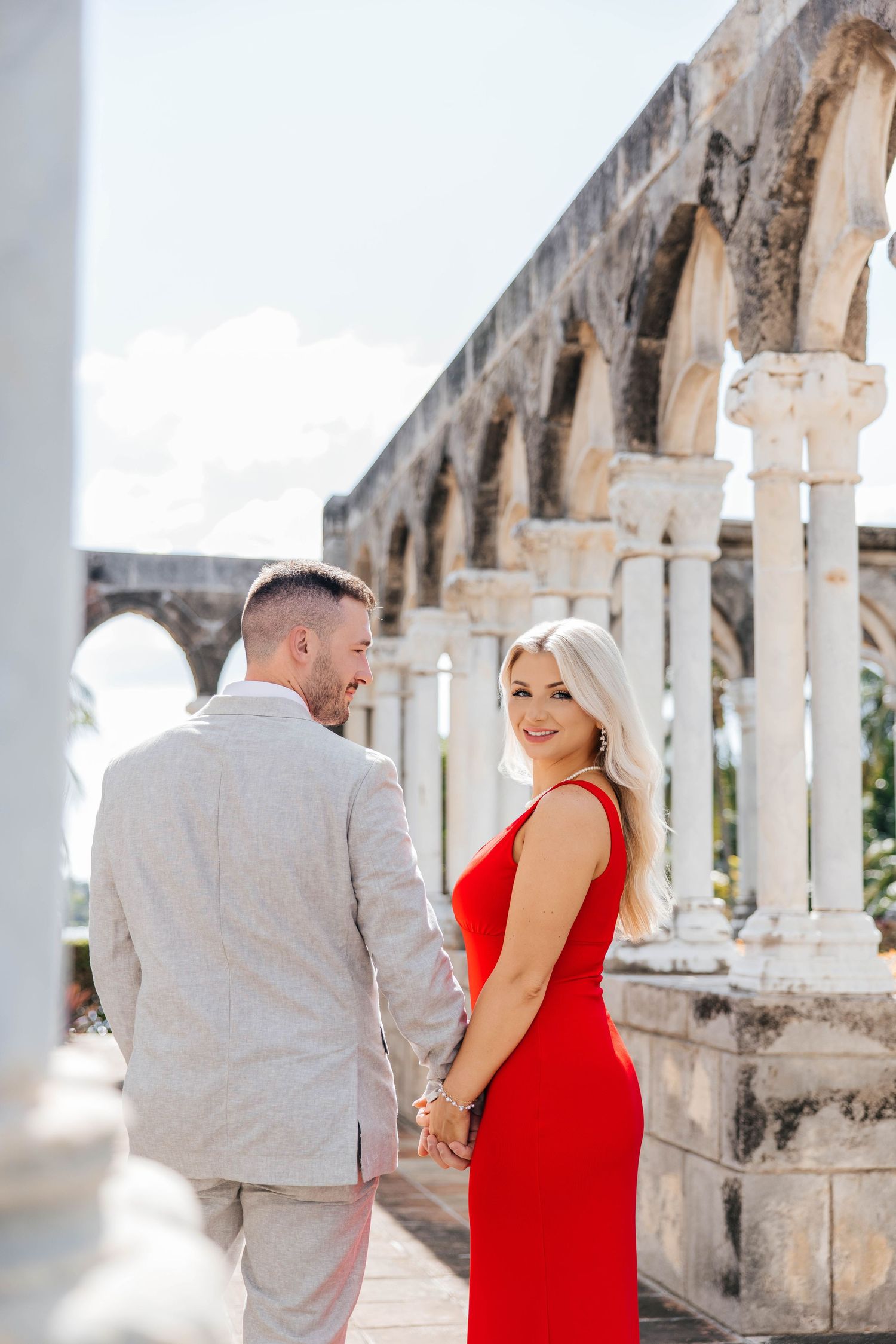 A couple in red and beige outfits poses together in front of ancient stone columns and archways.