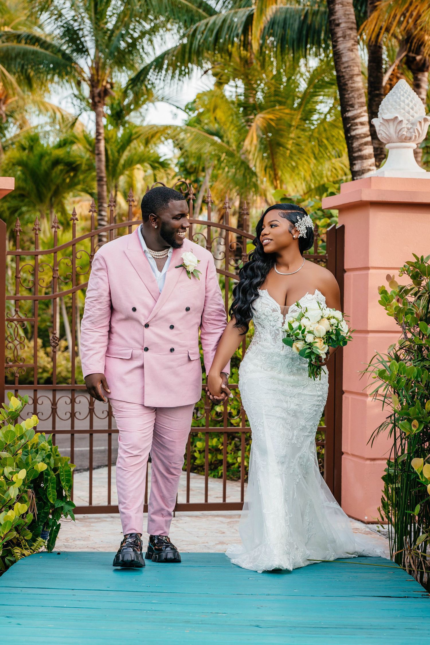 Wedding couple on turquoise bridge with pink suit and white gown against palm trees.