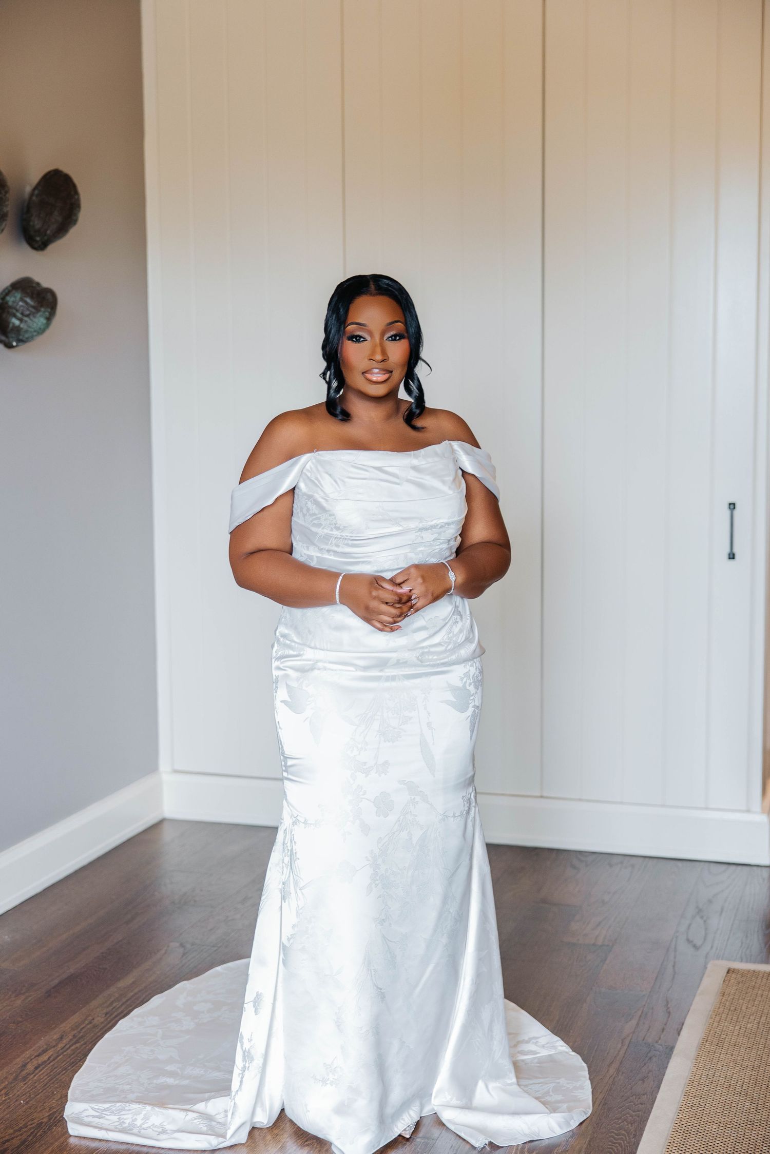 A bride in an elegant off-shoulder white satin mermaid wedding gown with a train poses indoors on hardwood floors.
