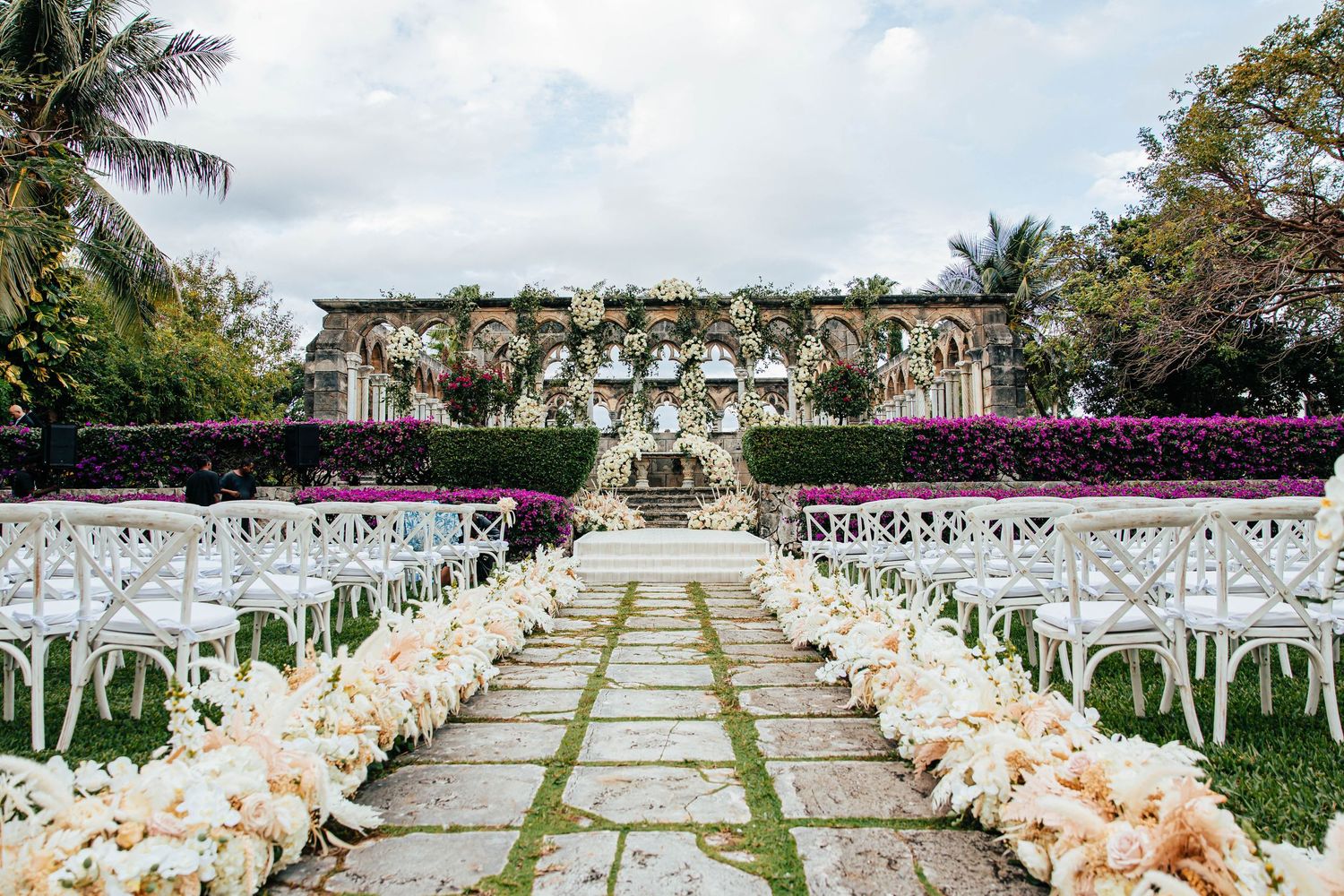 Elegant outdoor wedding ceremony aisle lined with white chairs and blush floral arrangements leading to a grand floral arch.