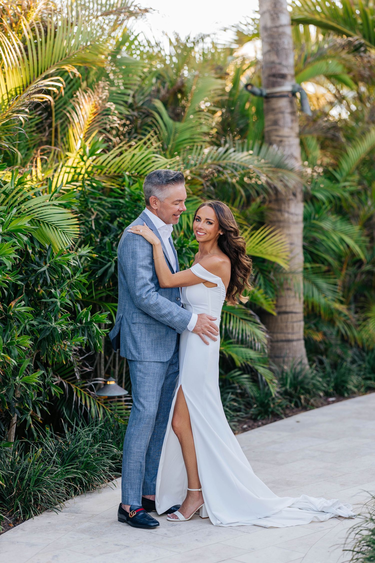 Couple embracing in tropical garden setting wearing elegant wedding attire with white dress and blue suit.