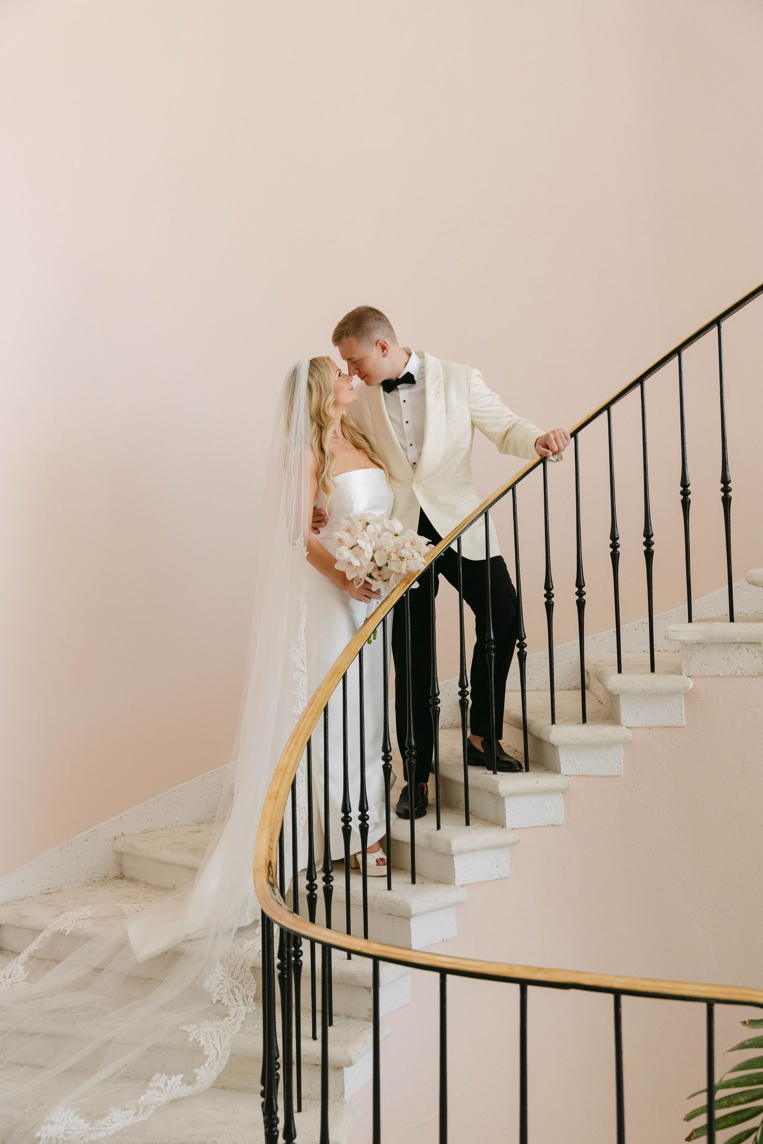 Bride in a strapless gown with a long lace-trimmed veil embraces her groom in a white tuxedo jacket on an elegant curved marble staircase.