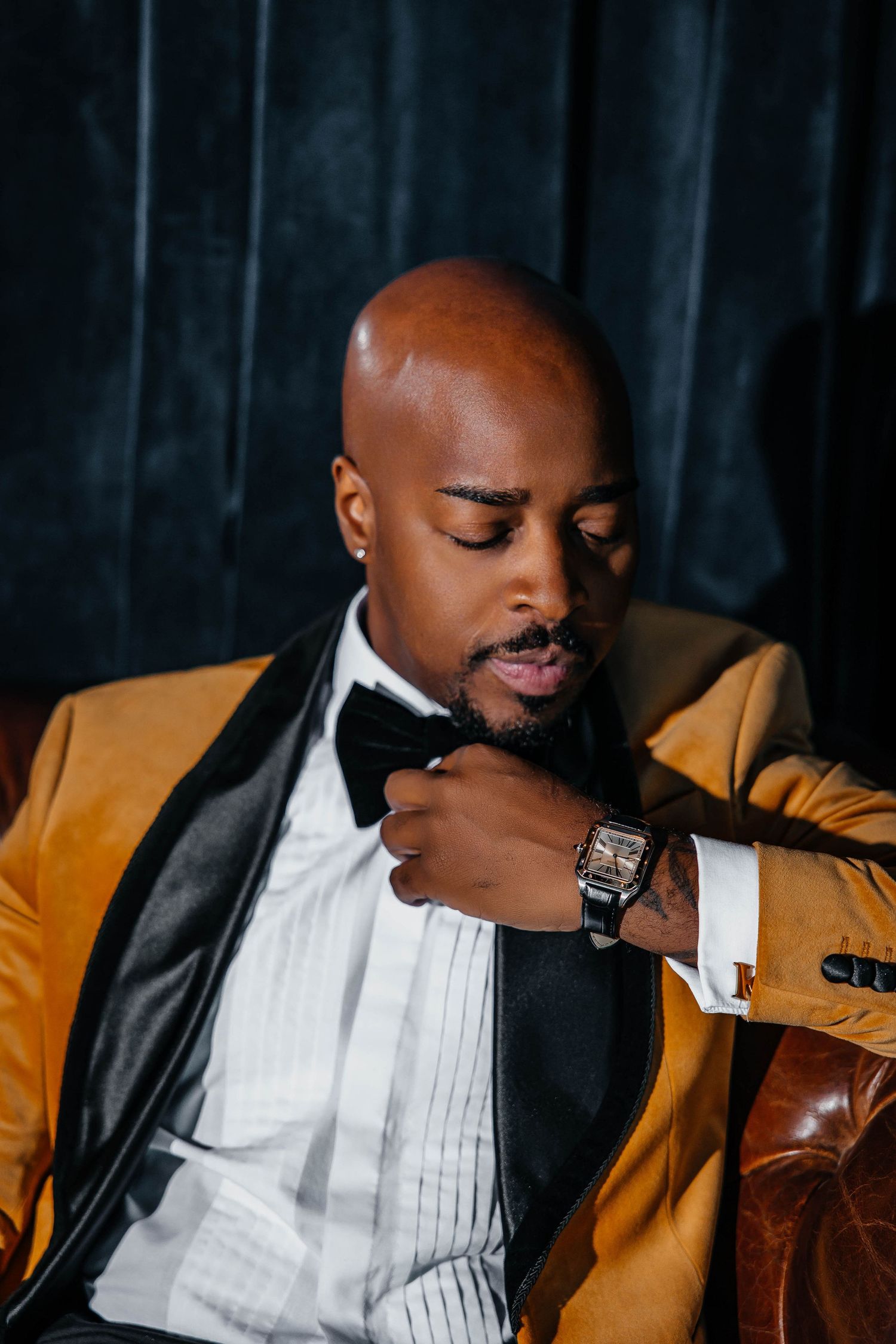 Sophisticated man in gold tuxedo jacket checks luxury watch against dark background.
