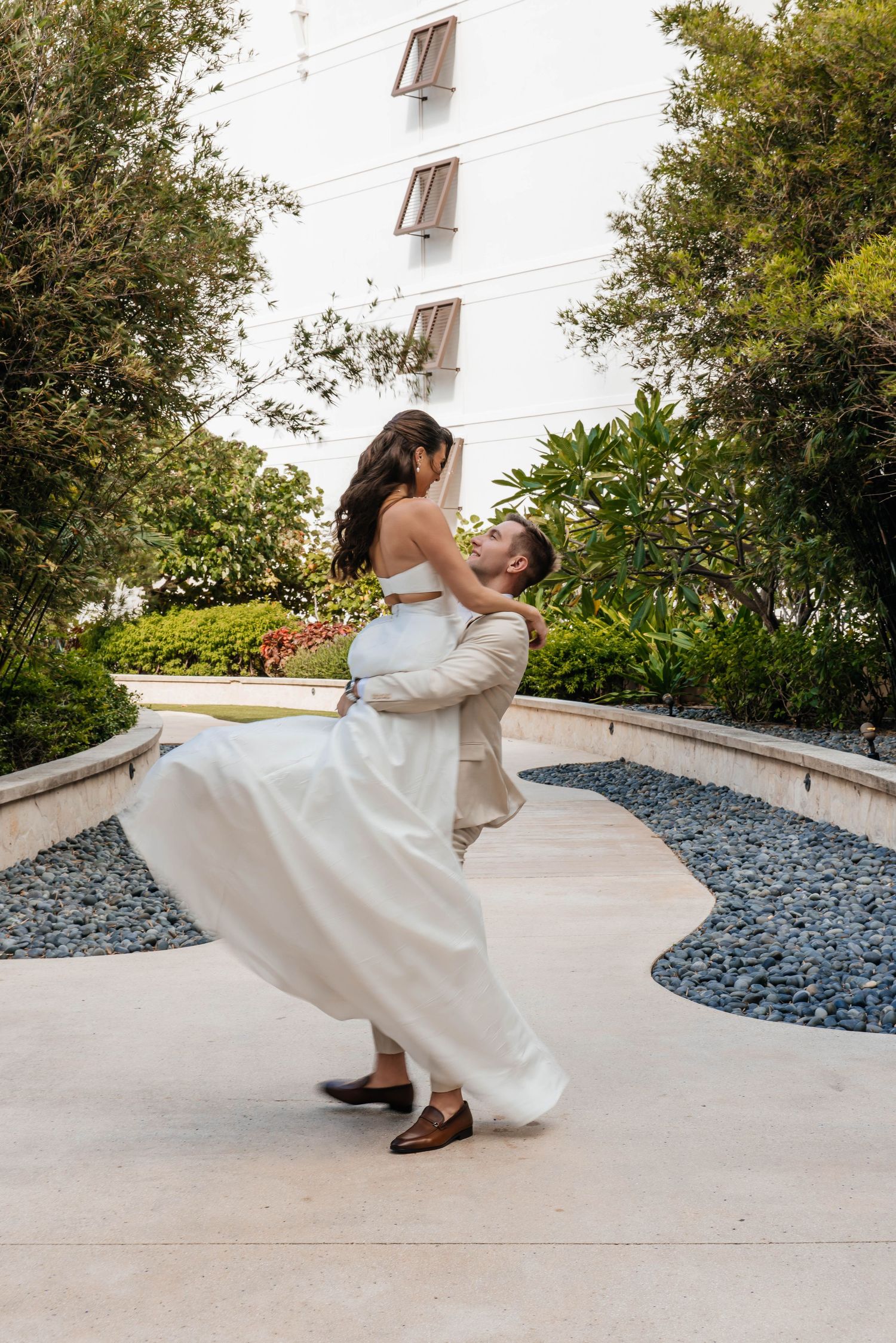 A couple performs a dramatic dip pose in front of a white building and landscaping on a concrete patio.