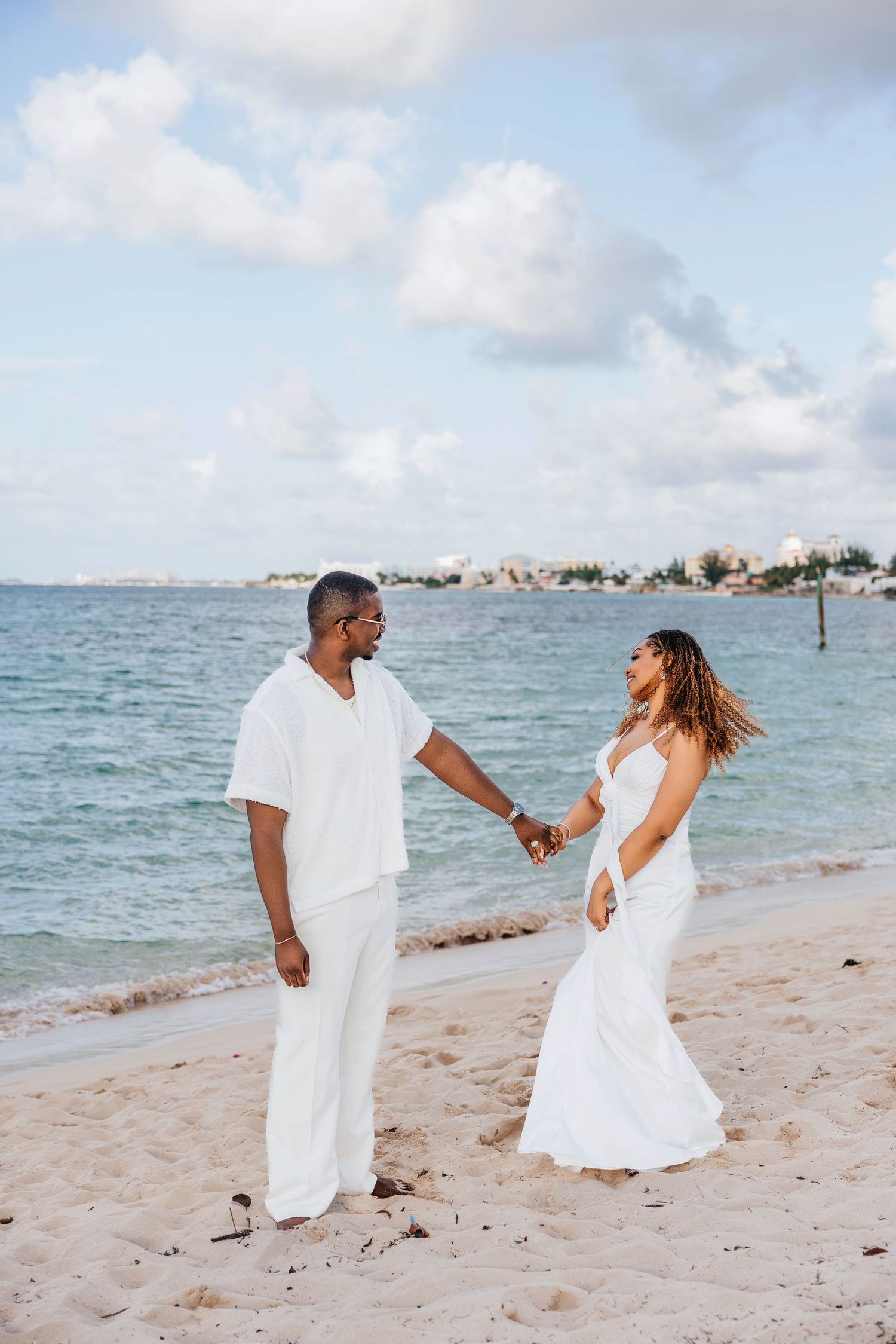A couple in all white outfits holds hands on a tropical beach with turquoise waters and blue skies in the background.