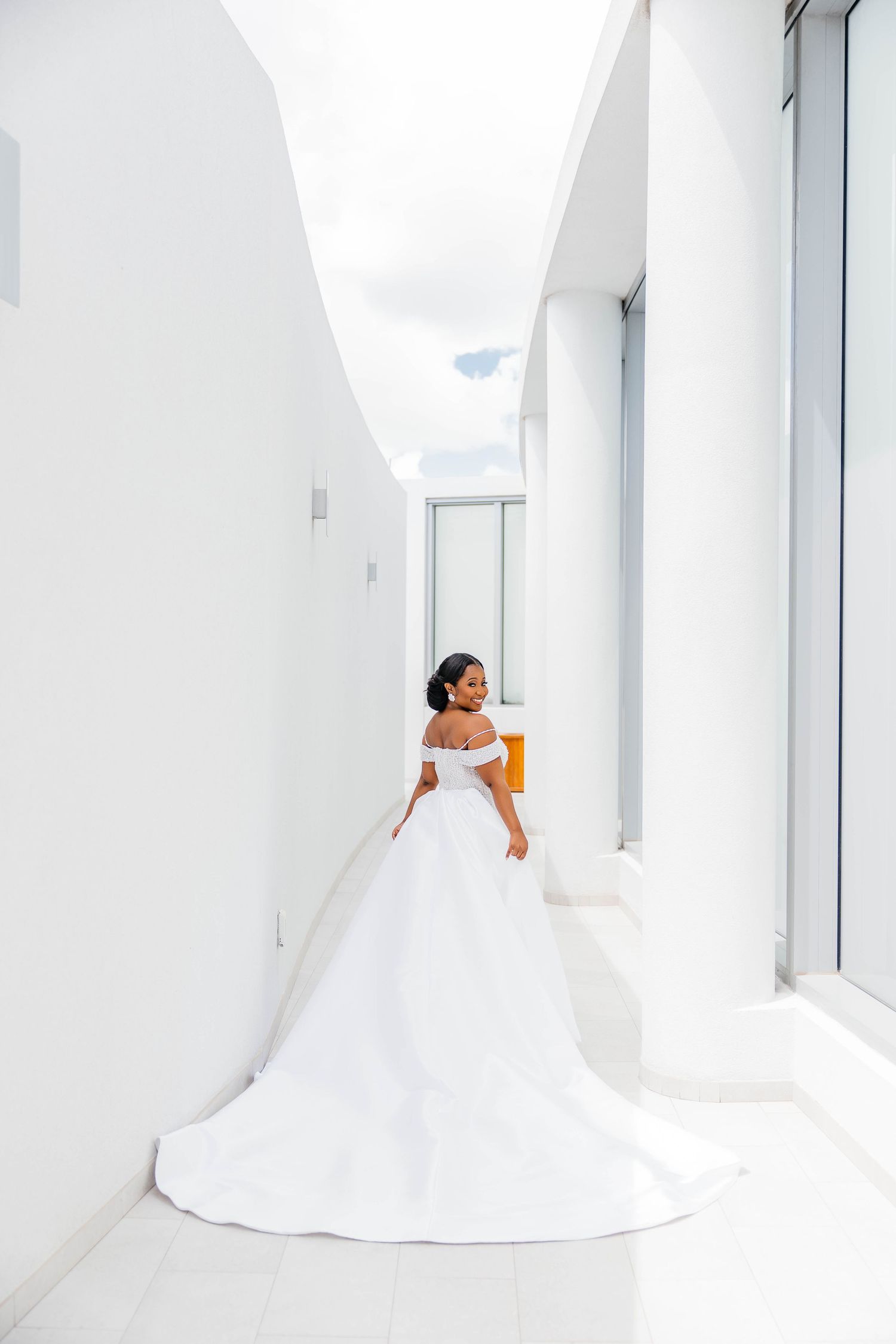 Bride in elegant off-shoulder white ball gown with long train poses in a bright modern white corridor.