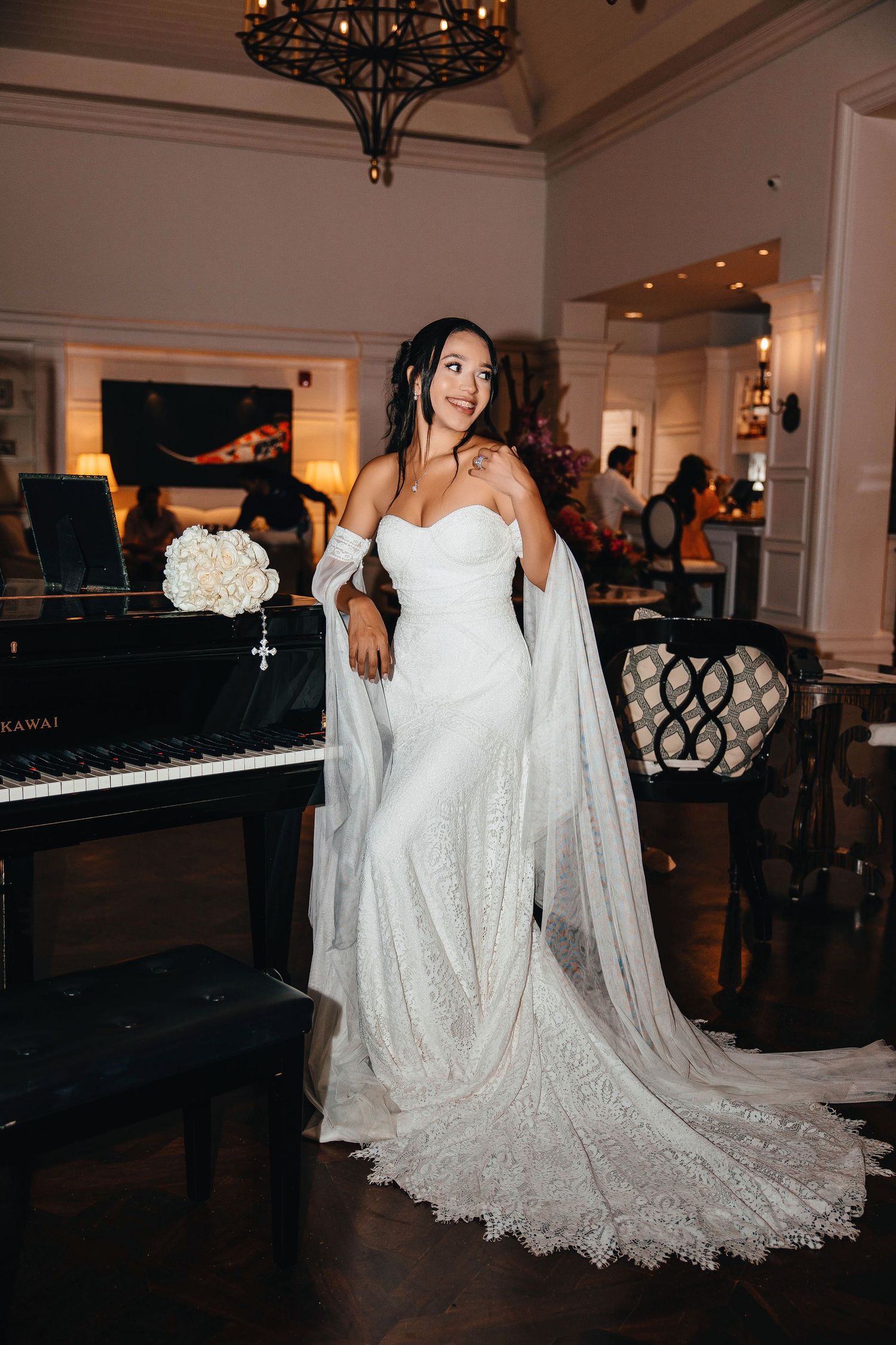 Bride in white lace strapless gown with long veil poses elegantly beside a black Kawai grand piano indoors.