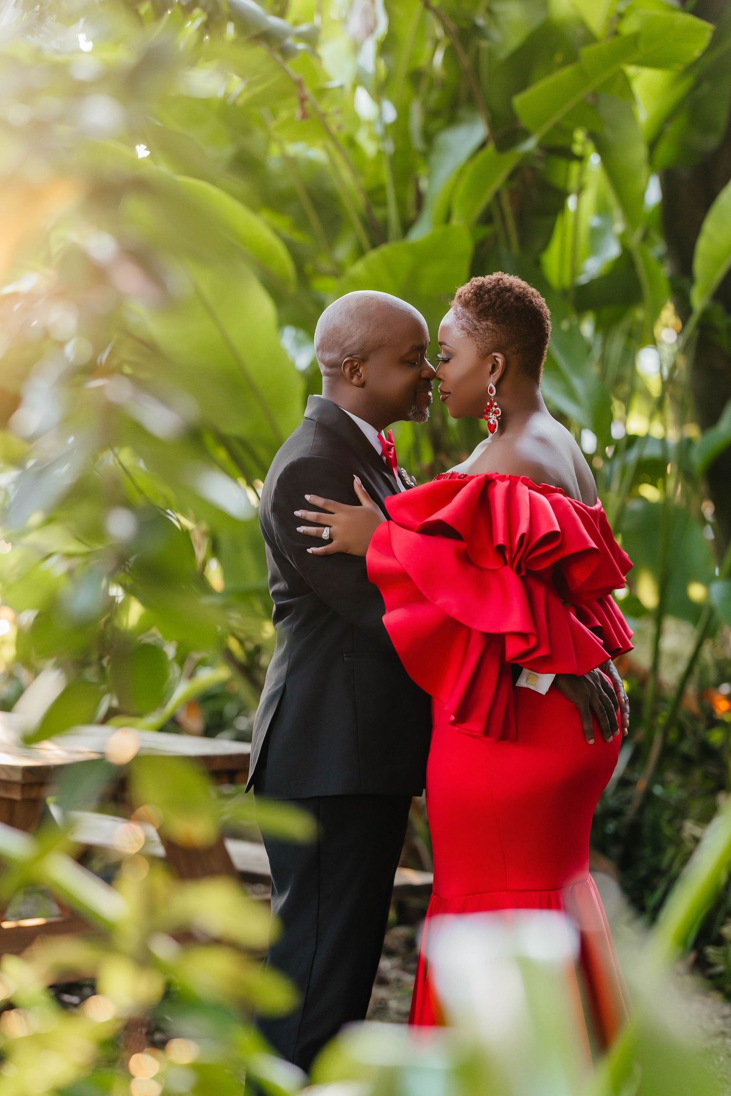 A couple embraces in a garden setting, with one wearing a dramatic red ruffled dress and the other in a black suit.