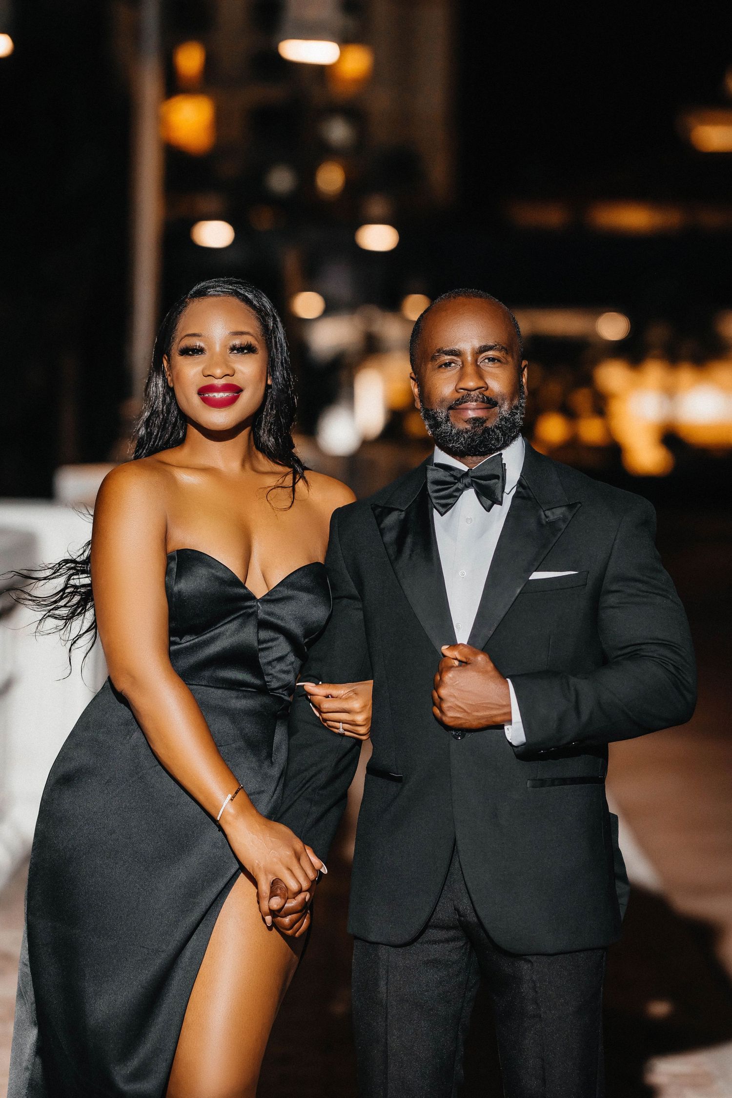 Stylish couple in formal black attire pose together for evening photoshoot.
