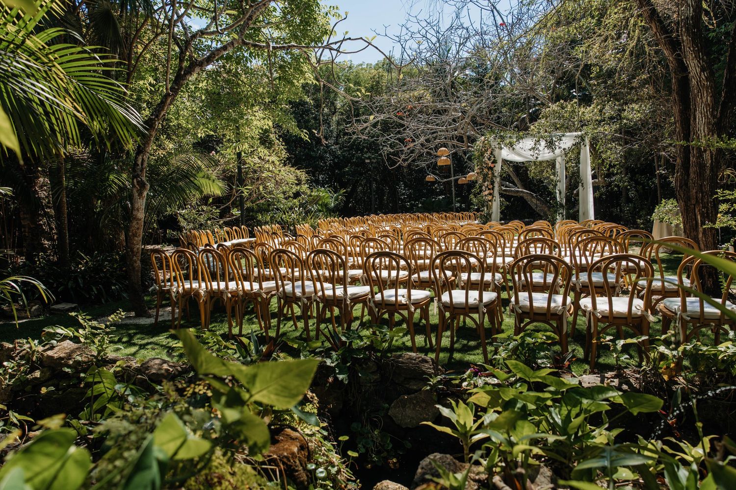Intimate outdoor wedding ceremony with wooden chairs set among tropical greenery and a white floral arch.