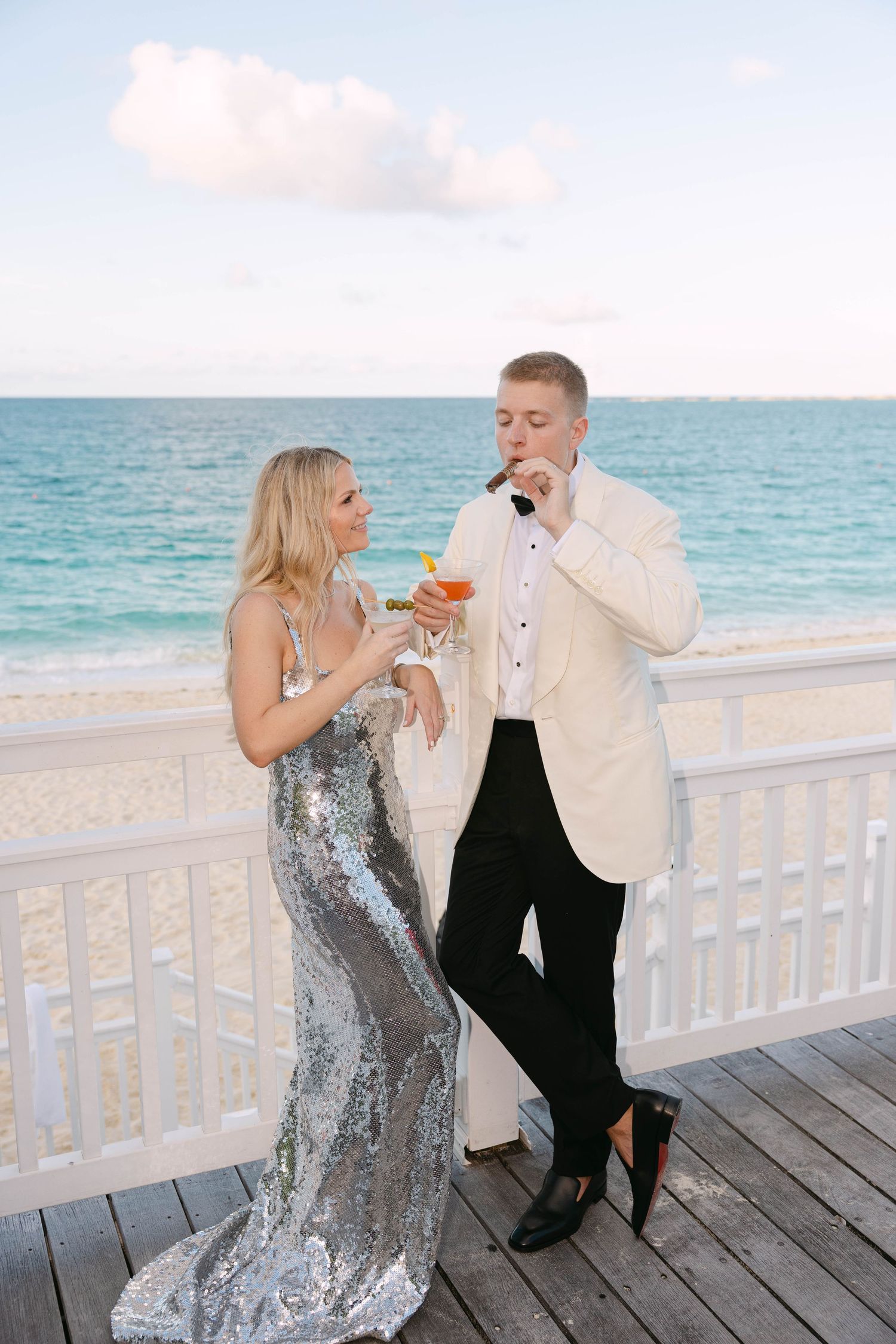 Elegant couple in a silver sequin gown and white dinner jacket toast with cocktails on a white beachfront deck overlooking turquoise ocean.