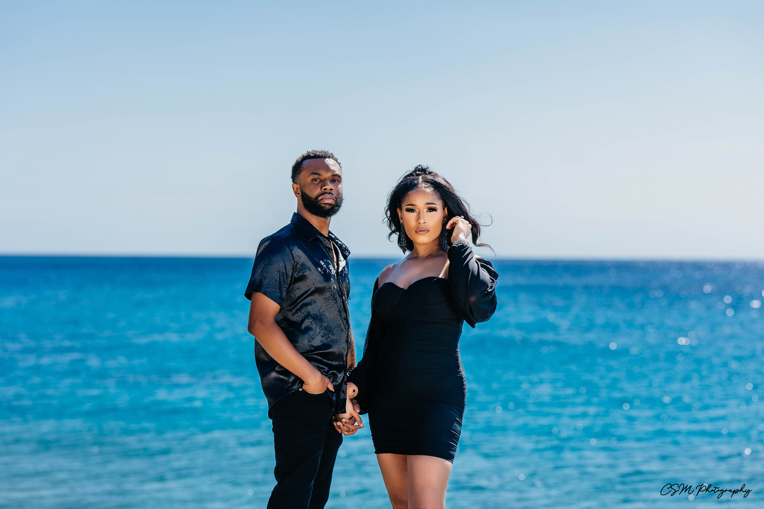 Stylish couple in black outfits posing on rocky coastline with bright blue ocean in background.