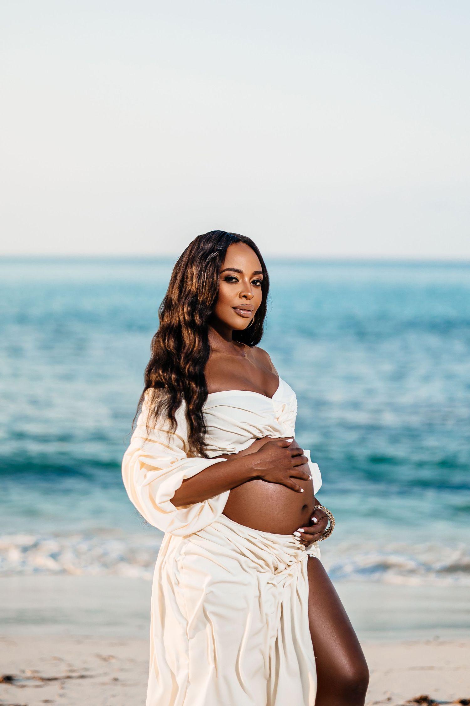 Maternity beach portrait with flowing white dress against ocean backdrop.