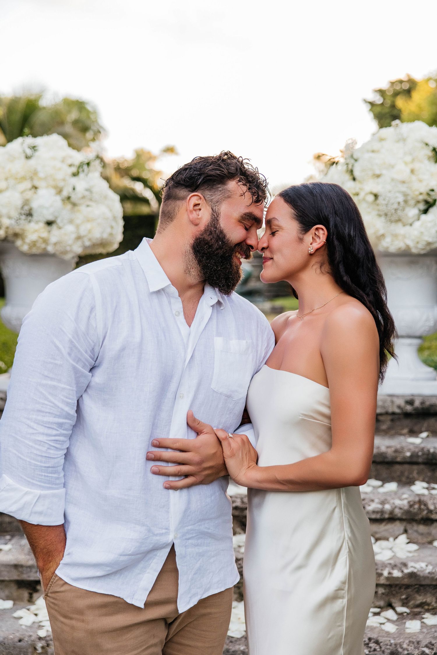 A couple shares an intimate moment in front of white floral arrangements outdoors.