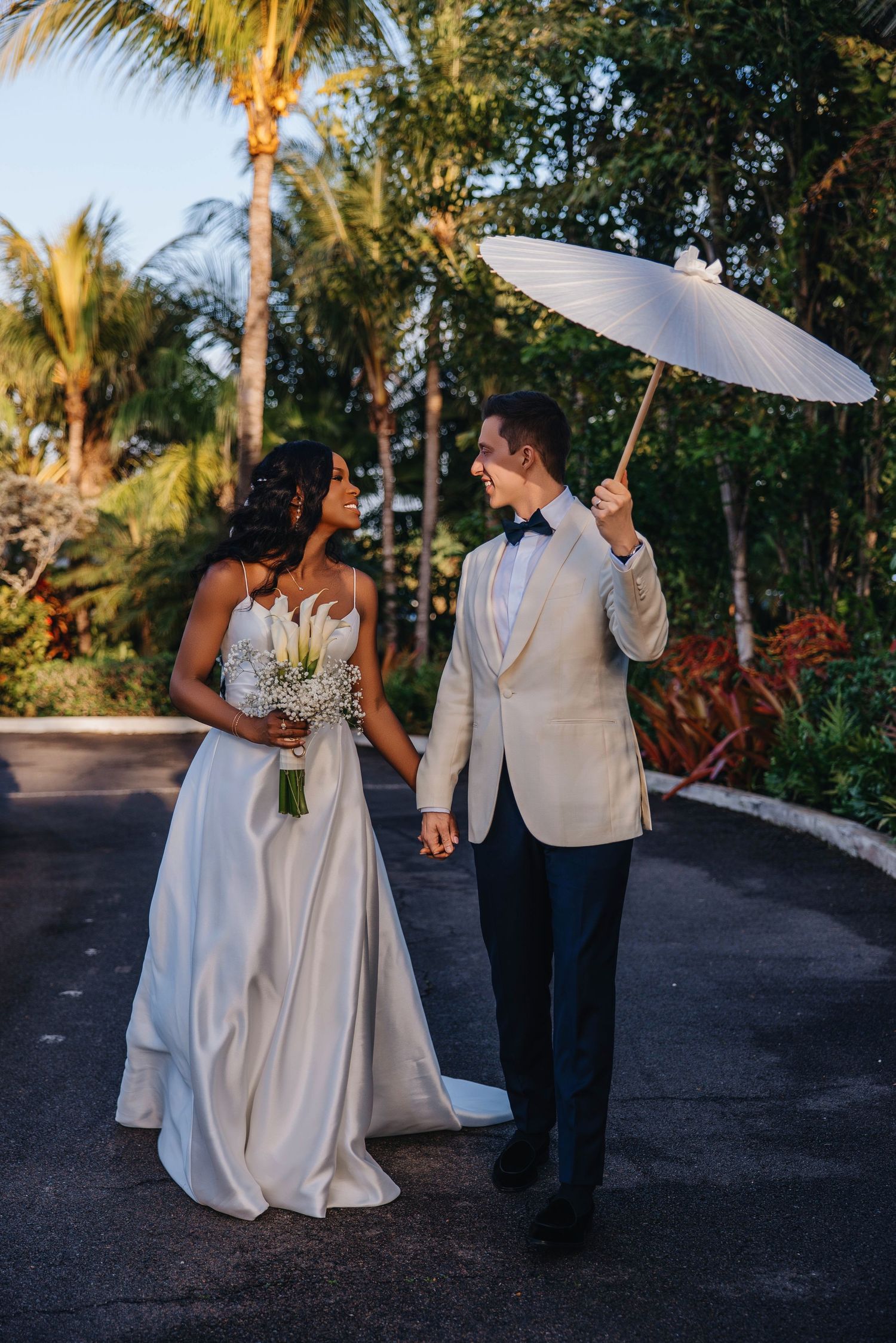 Bride and groom walking with parasol on tropical garden path during sunset wedding.