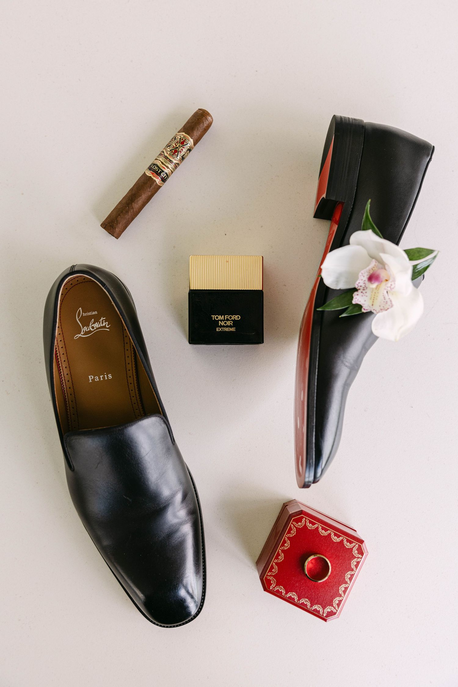 Black Christian Louboutin loafers, Tom Ford Noir Extreme cologne, a cigar, an orchid boutonniere and a red Cartier ring box flat lay.