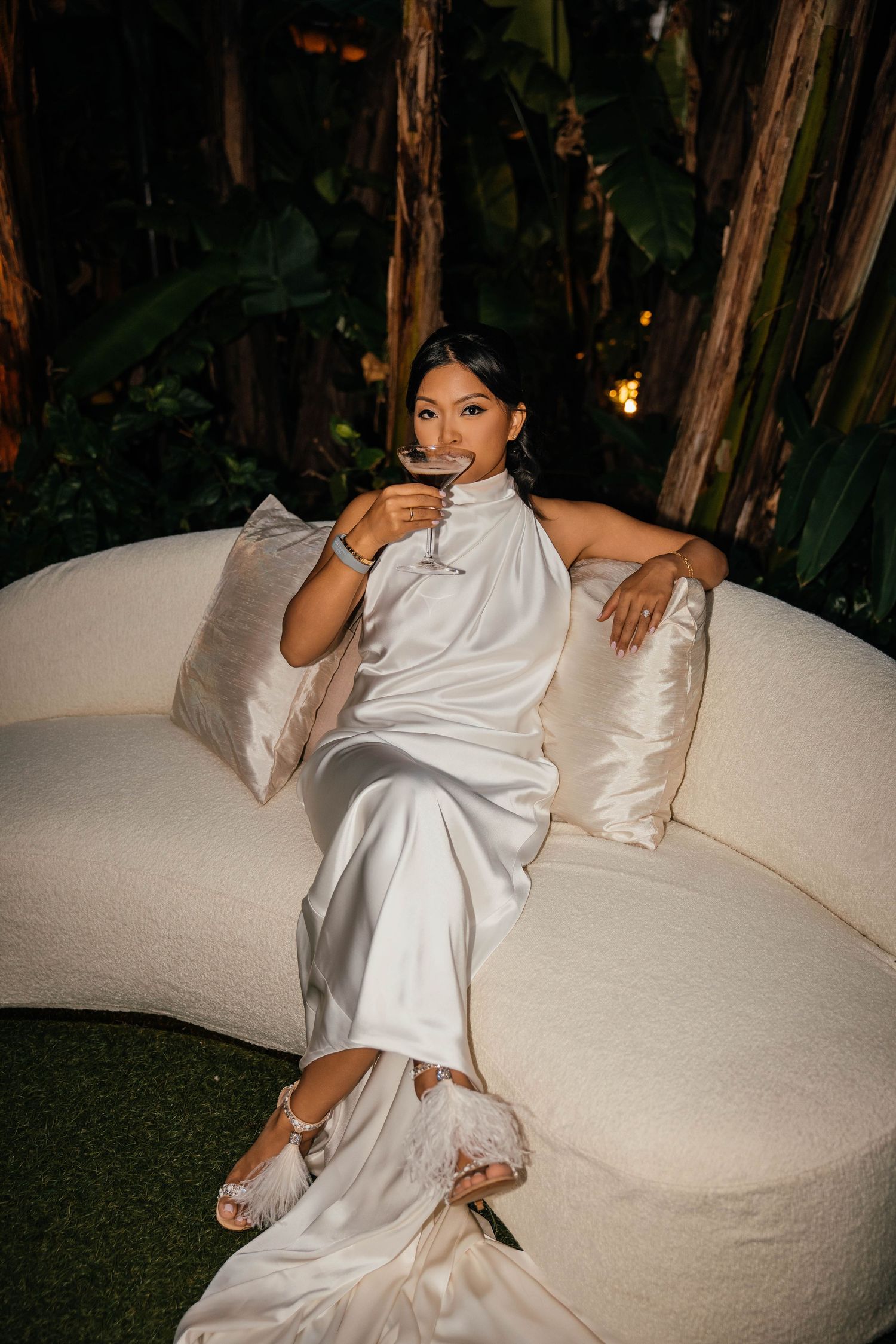 Woman in white satin dress sipping red wine on curved ivory sofa, surrounded by lush tropical greenery at night.