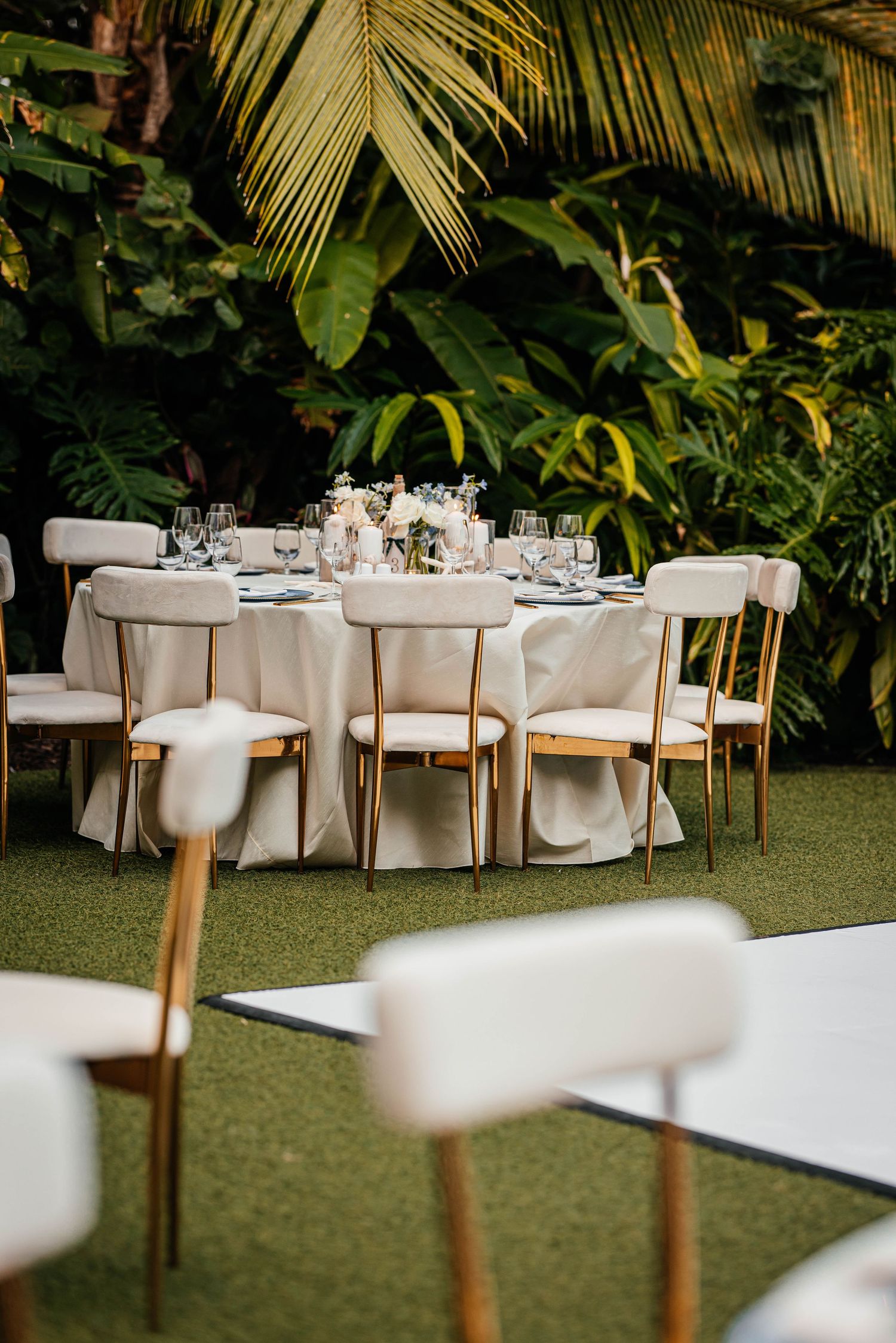 Elegant outdoor wedding reception with white chairs and round tables set against lush tropical greenery.
