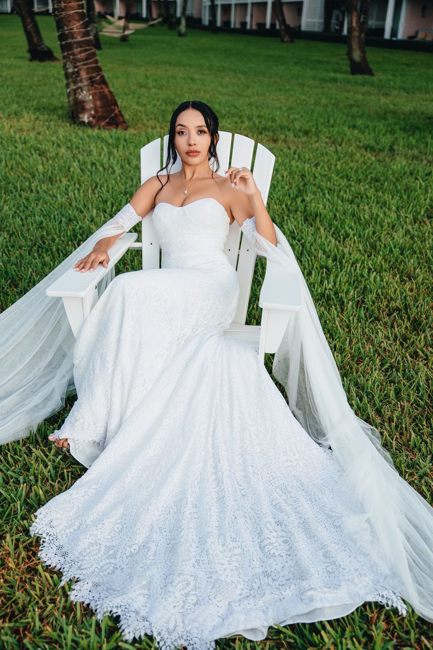 Bride in white lace mermaid gown sitting in Adirondack chair on green lawn.