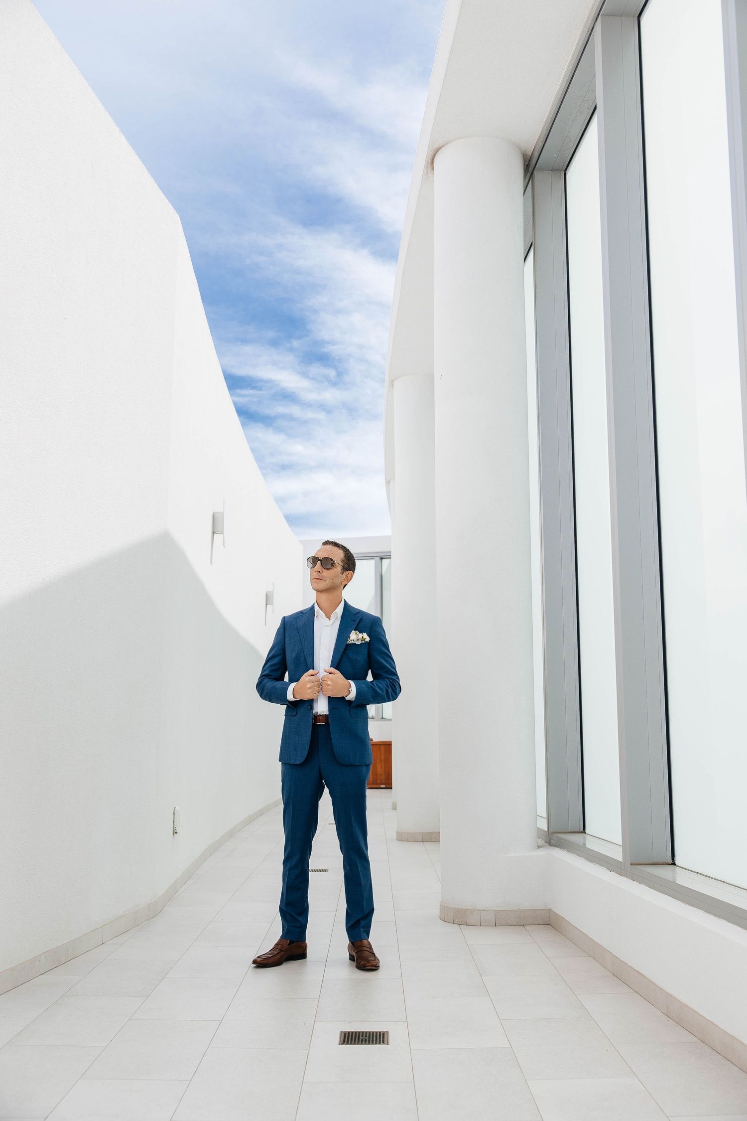 A well-dressed groom in a blue suit and sunglasses stands confidently on a modern white rooftop terrace.