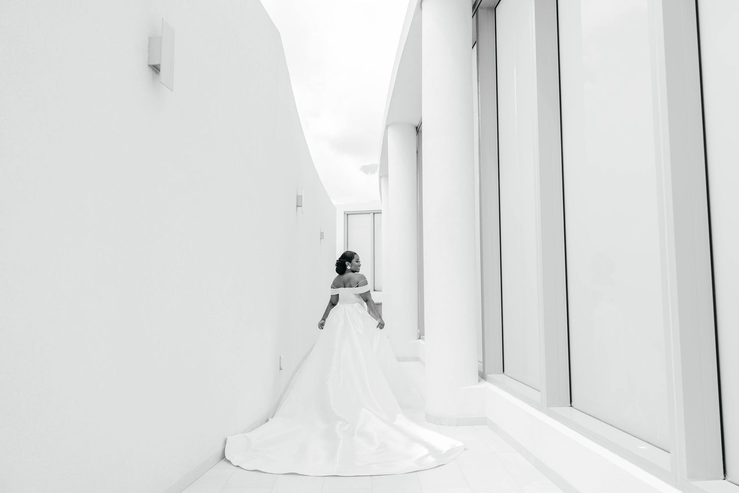 Bride in a flowing white gown stands in a minimalist white corridor, captured in elegant black and white photography.