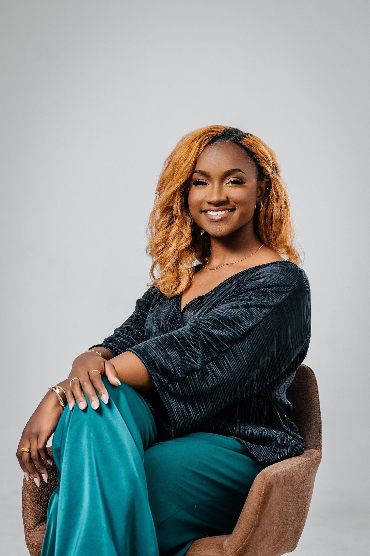 Professional studio portrait of a person in a navy top and teal pants relaxing in a brown chair.