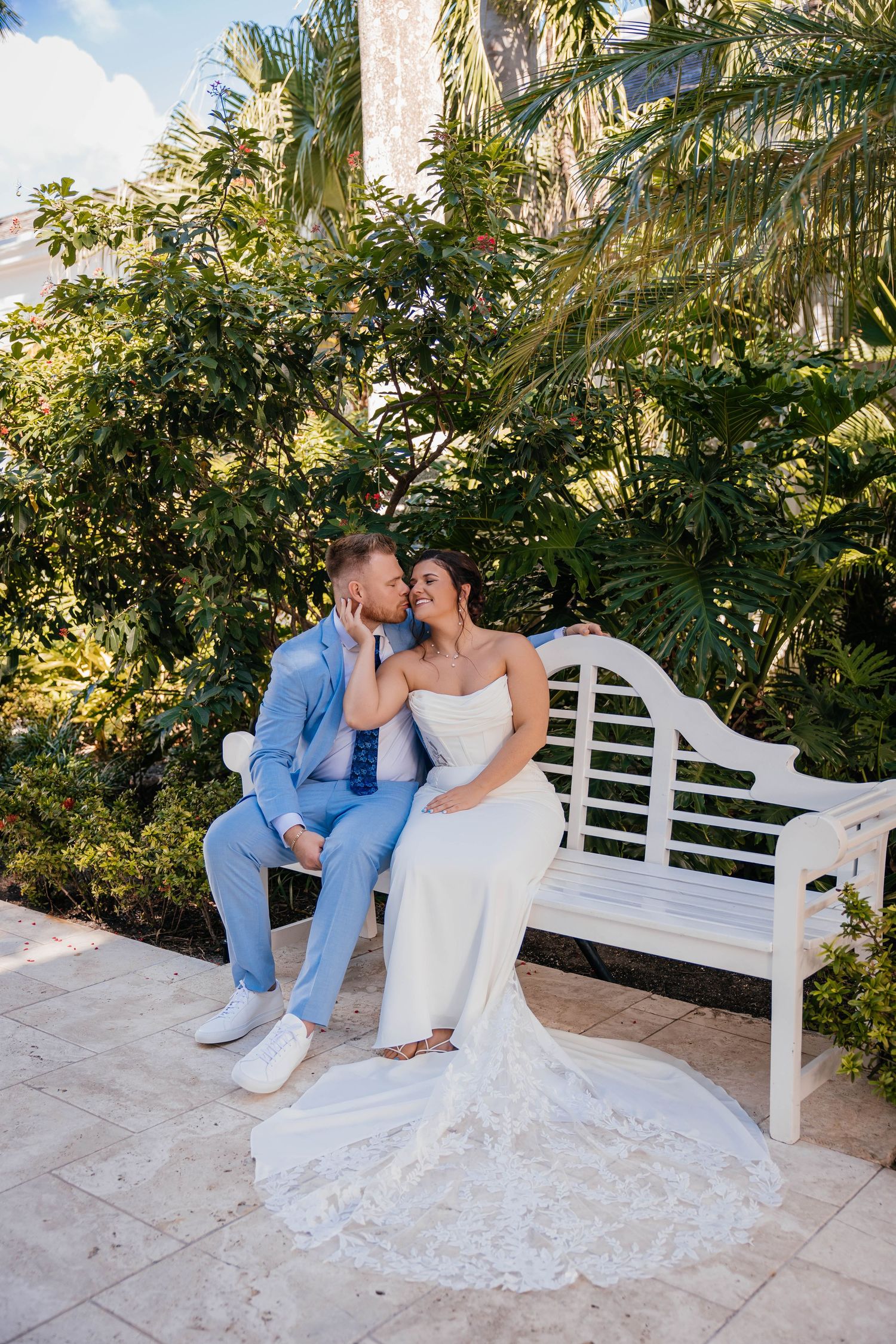 A romantic couple sits on a white garden bench surrounded by lush palm trees and greenery in a tropical setting.