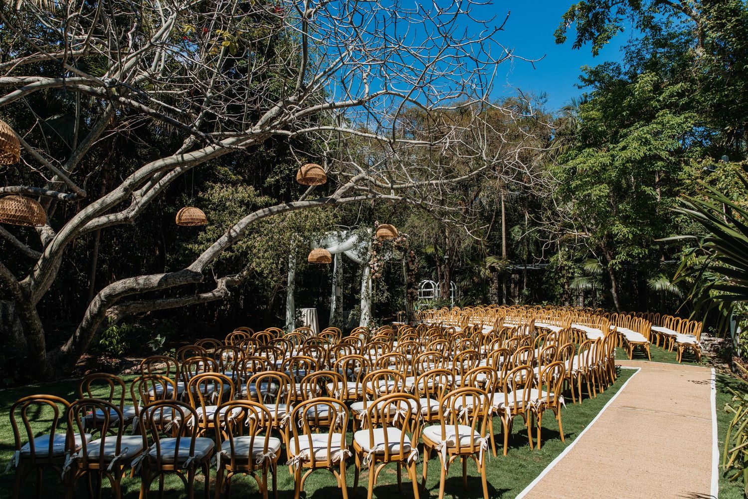 Outdoor wedding ceremony setup with wooden chairs arranged in rows on a lush garden path beneath a large tree.