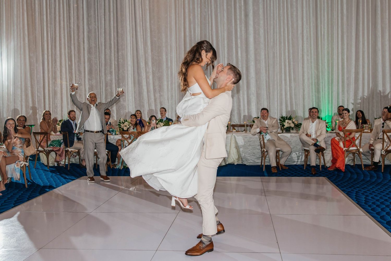 Groom lifts bride during first dance at elegant wedding reception with white draping and guests watching.