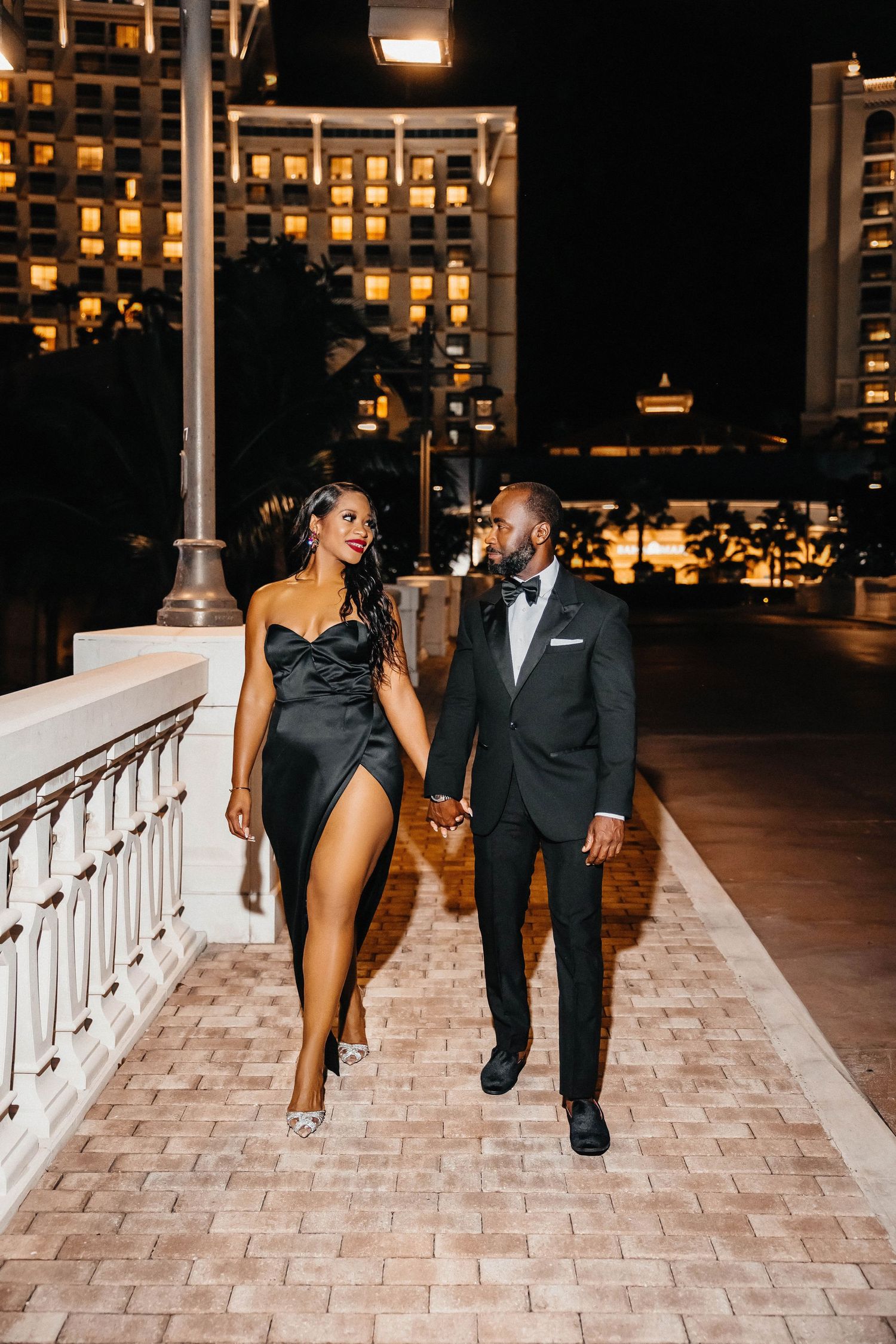 Glamorous couple walks hand in hand on illuminated balcony at night.
