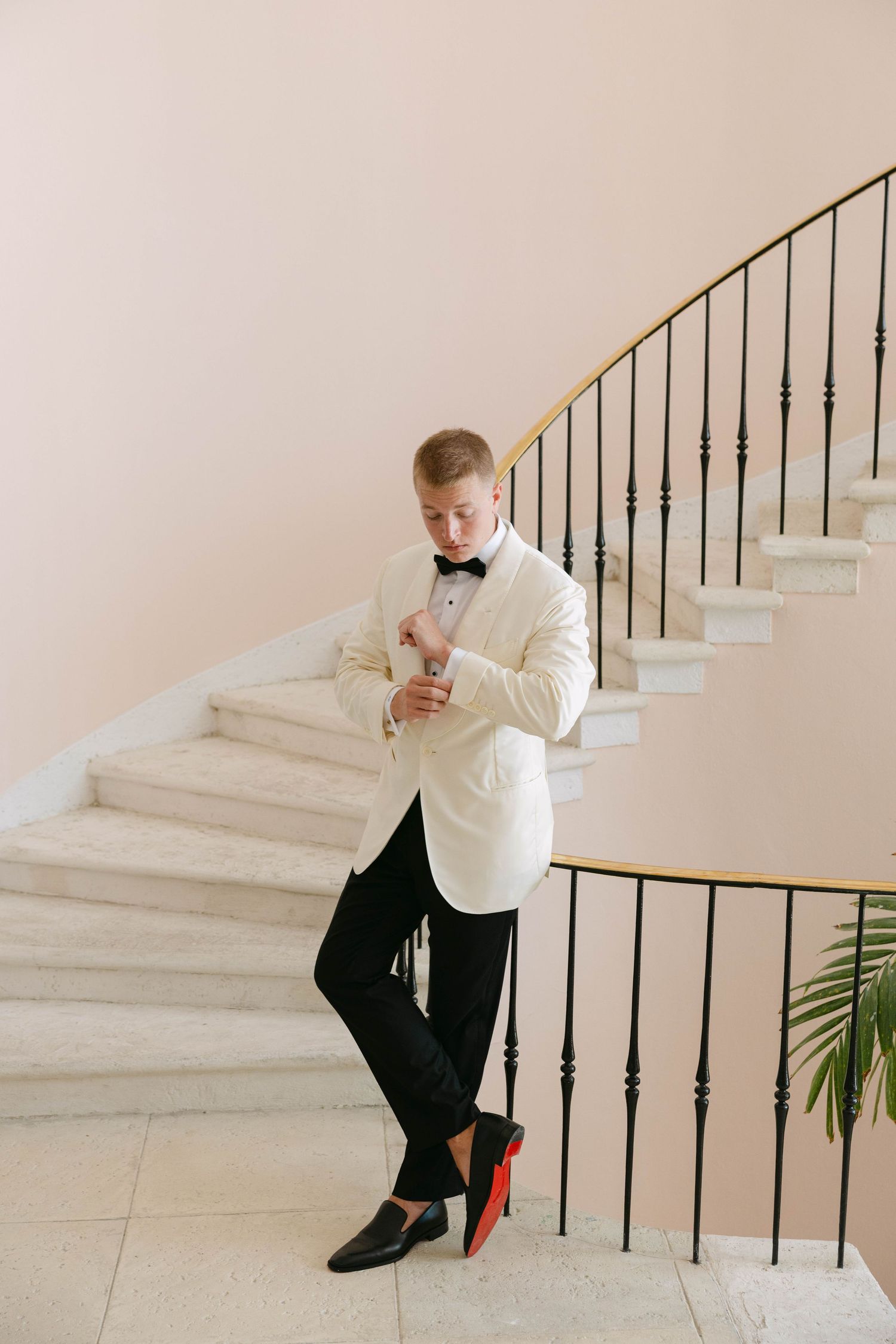 Groom in a white dinner jacket, black bow tie and red-soled loafers adjusts his cufflink on a curved staircase with a wrought iron railing.