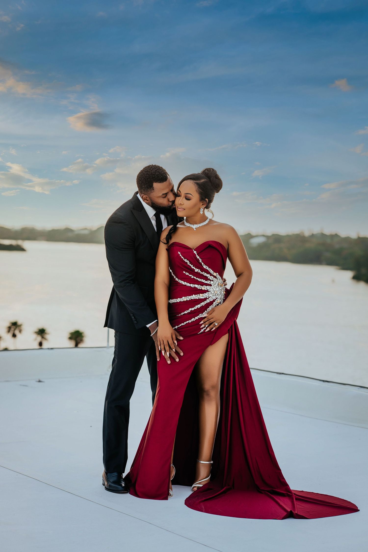 Elegant couple poses on rooftop at sunset, woman wearing stunning burgundy gown with crystal details and dramatic train.