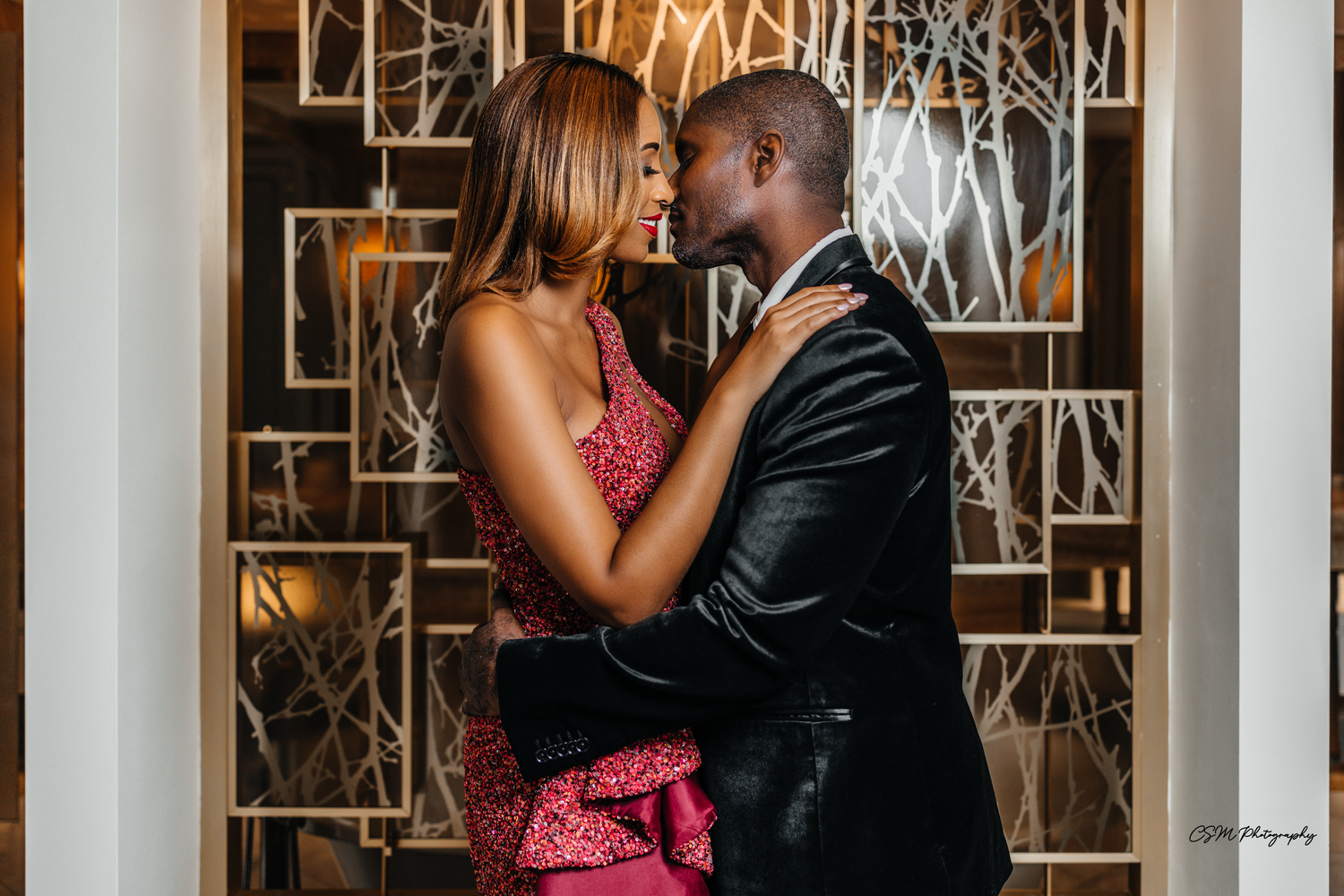 Romantic couple in formal wear shares intimate moment against decorative wall.