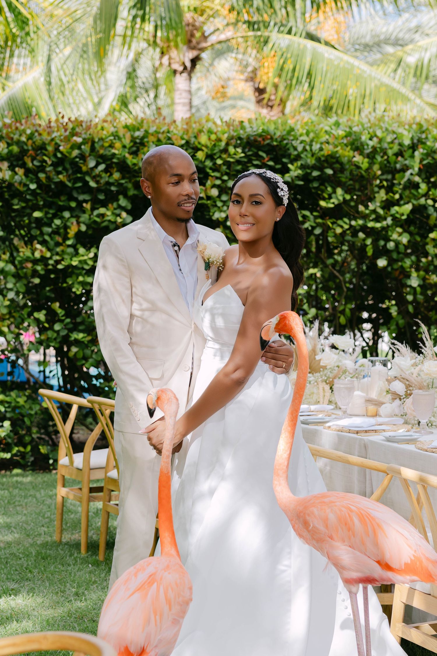 A bride and groom pose with pink flamingos at an outdoor tropical wedding reception with palm trees and elegant table settings.