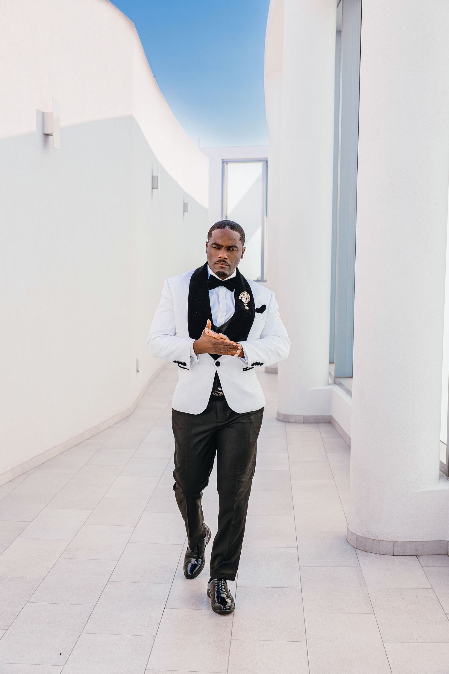 Elegant man in white tuxedo jacket and black bow tie walking confidently through a modern white corridor.