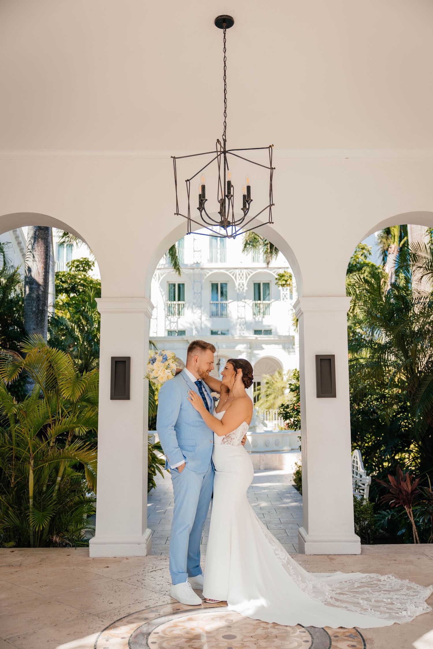 Romantic wedding couple shares intimate moments under elegant white archways with tropical foliage in background.
