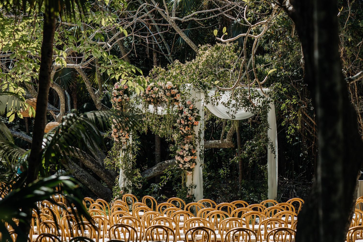 Outdoor wedding ceremony setup with rattan chairs and floral arch amid lush tropical greenery.