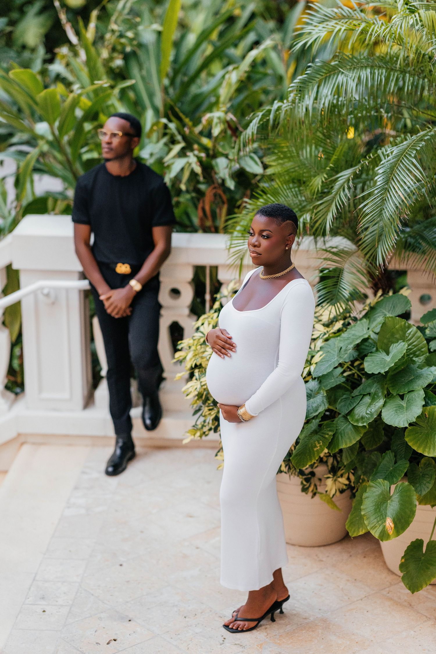 Expecting couple poses by lush tropical plants during outdoor maternity session.