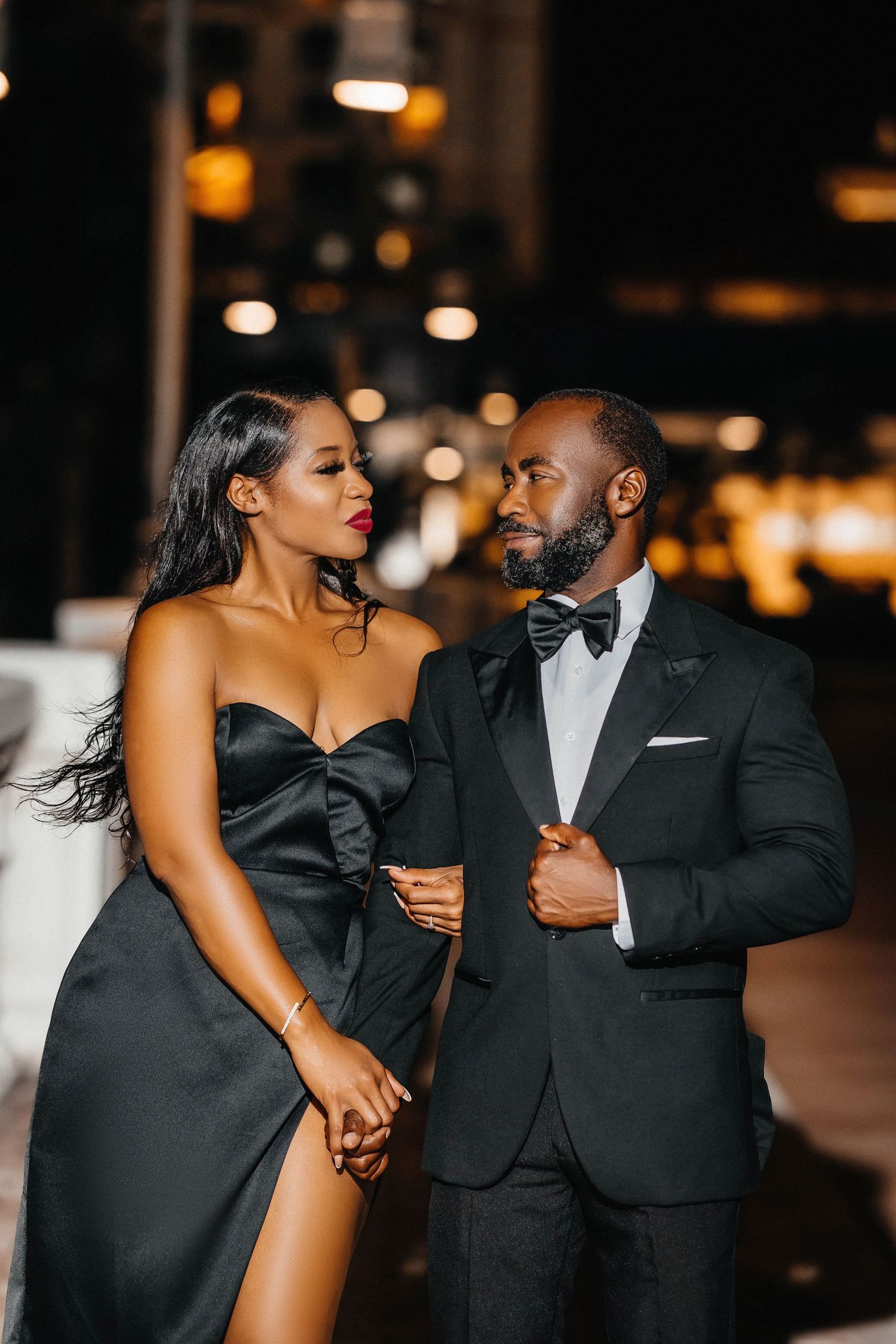 Elegant couple in black formal wear pose together against city lights at night.